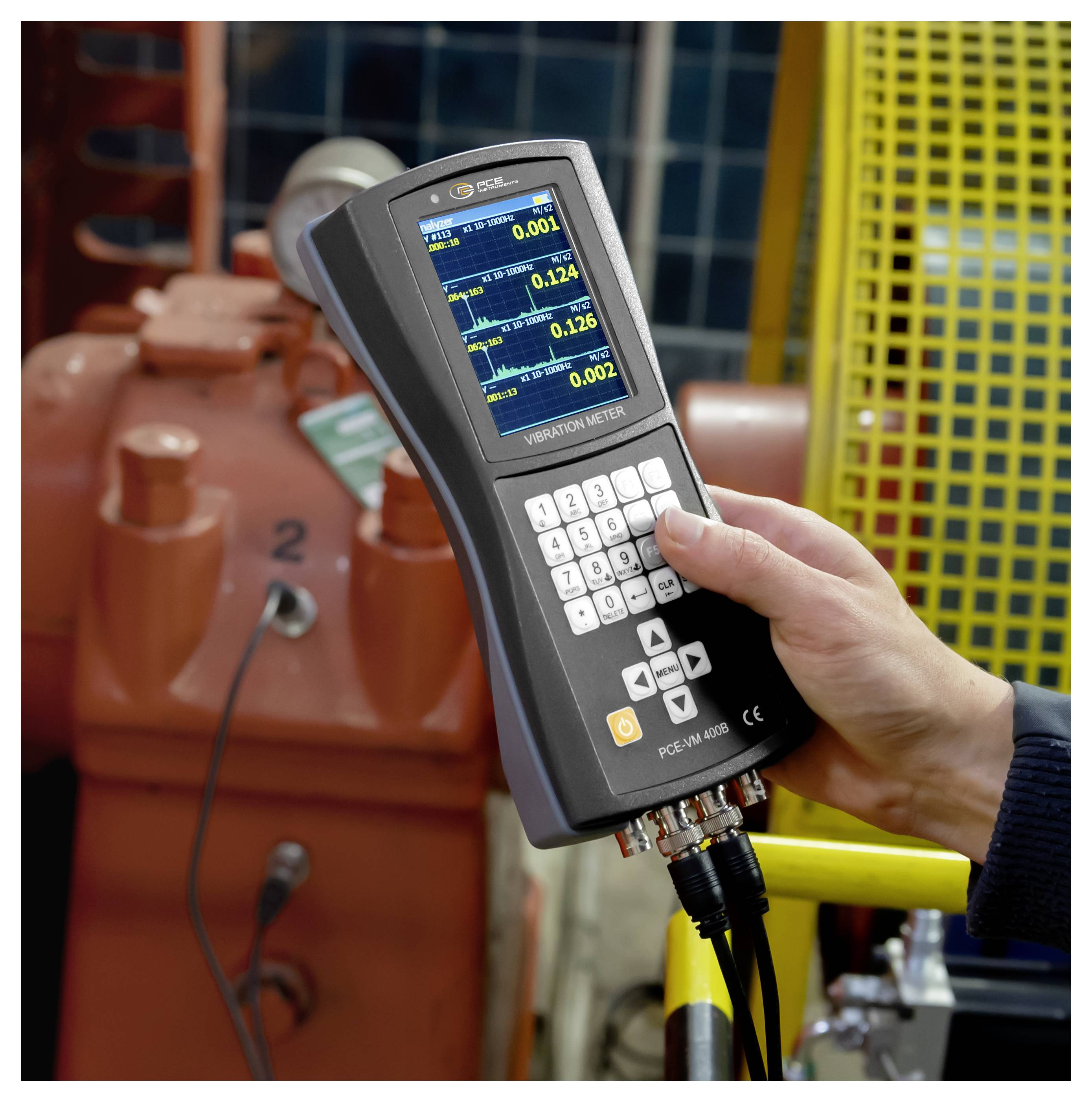 Hand die een trillingmeter vasthoudt met live-aflezingen, meetapparatuur in een industriële omgeving in de buurt van een machine of motor.
