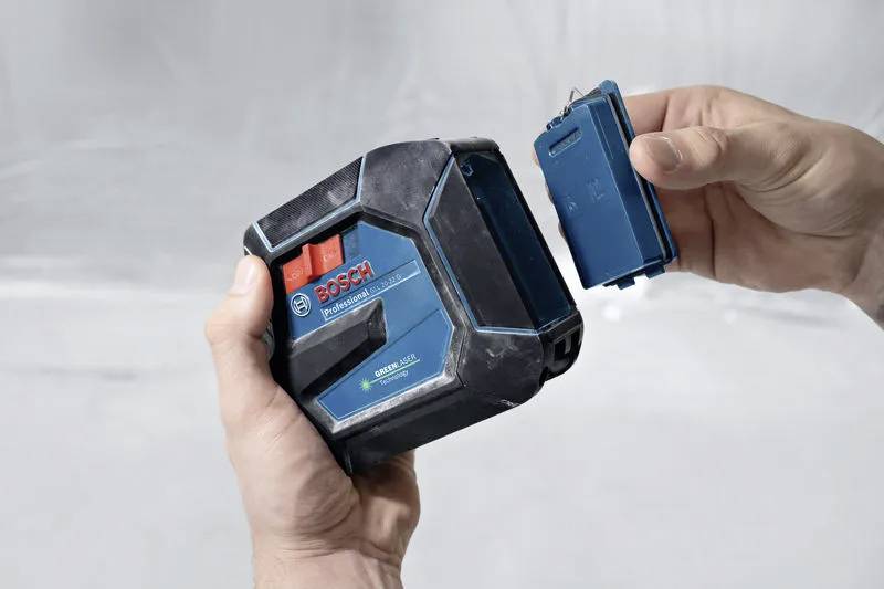 Een persoon houdt een Bosch lasermeetinstrument vast en plaatst een klein blauw verwijderbaar onderdeel. De achtergrond is een neutrale grijze ondergrond.