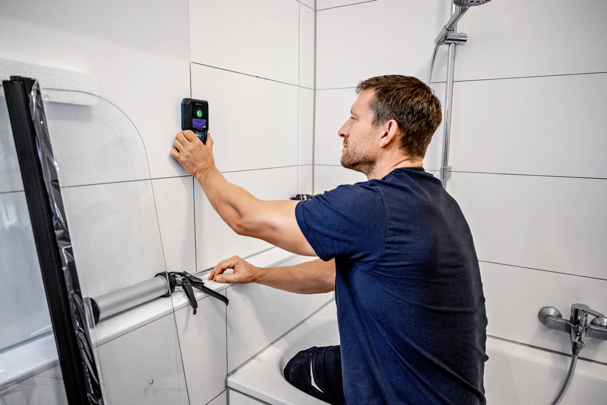 Een man knielt in een badkuip en meet de tegels in de badkamer met een digitaal apparaat.