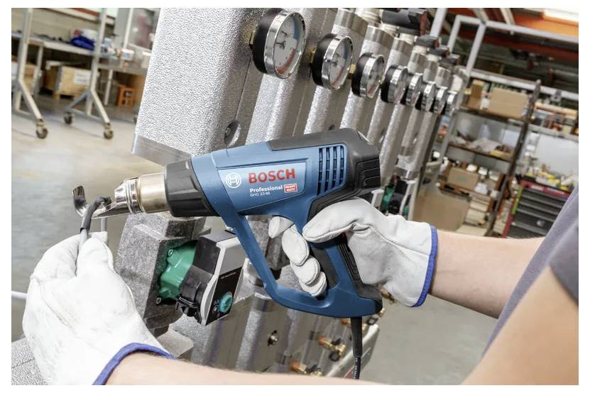 Een persoon met handschoenen gebruikt een Bosch heteluchtpistool op buisisolatie, met industriële meters op de achtergrond, in een werkplaatsomgeving.
