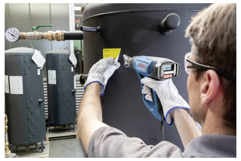 Een persoon met handschoenen gebruikt een heteluchtpistool om een geel label op een grote industriële tank aan te brengen, met andere tanks zichtbaar op de achtergrond.