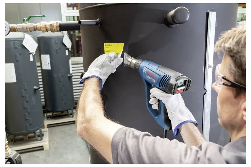 Een persoon die een heteluchtpistool gebruikt op een geel etiket dat is aangebracht op een grote cilindrische tank, terwijl hij handschoenen en beschermende oogapparatuur draagt in een industriële omgeving.