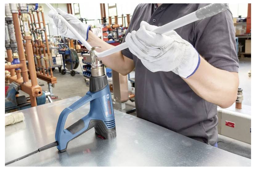 Een persoon met handschoenen gebruikt een blauwe heteluchtpistool om een grijze kunststof pijp te buigen op een werkbanktafel, met verschillende gereedschappen op de achtergrond.