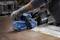 Een persoon die een blauwe Bosch schuurmachine gebruikt op een geroeste metalen oppervlakte, terwijl hij beschermende handschoenen draagt.