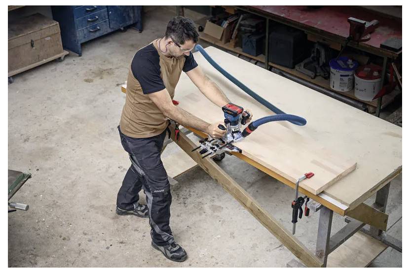 Een persoon gebruikt een elektrisch gereedschap om te werken aan een groot stuk hout in een werkplaats. Werkbanken en gereedschappen zijn zichtbaar op de achtergrond.