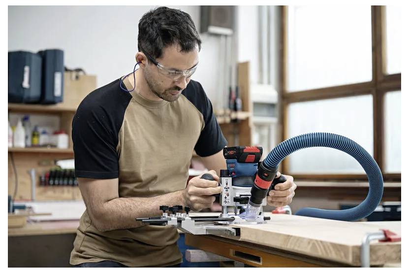 Een man met veiligheidsbril en oordopjes bedient een elektrisch gereedschap op een houten werkbank in een werkplaats.