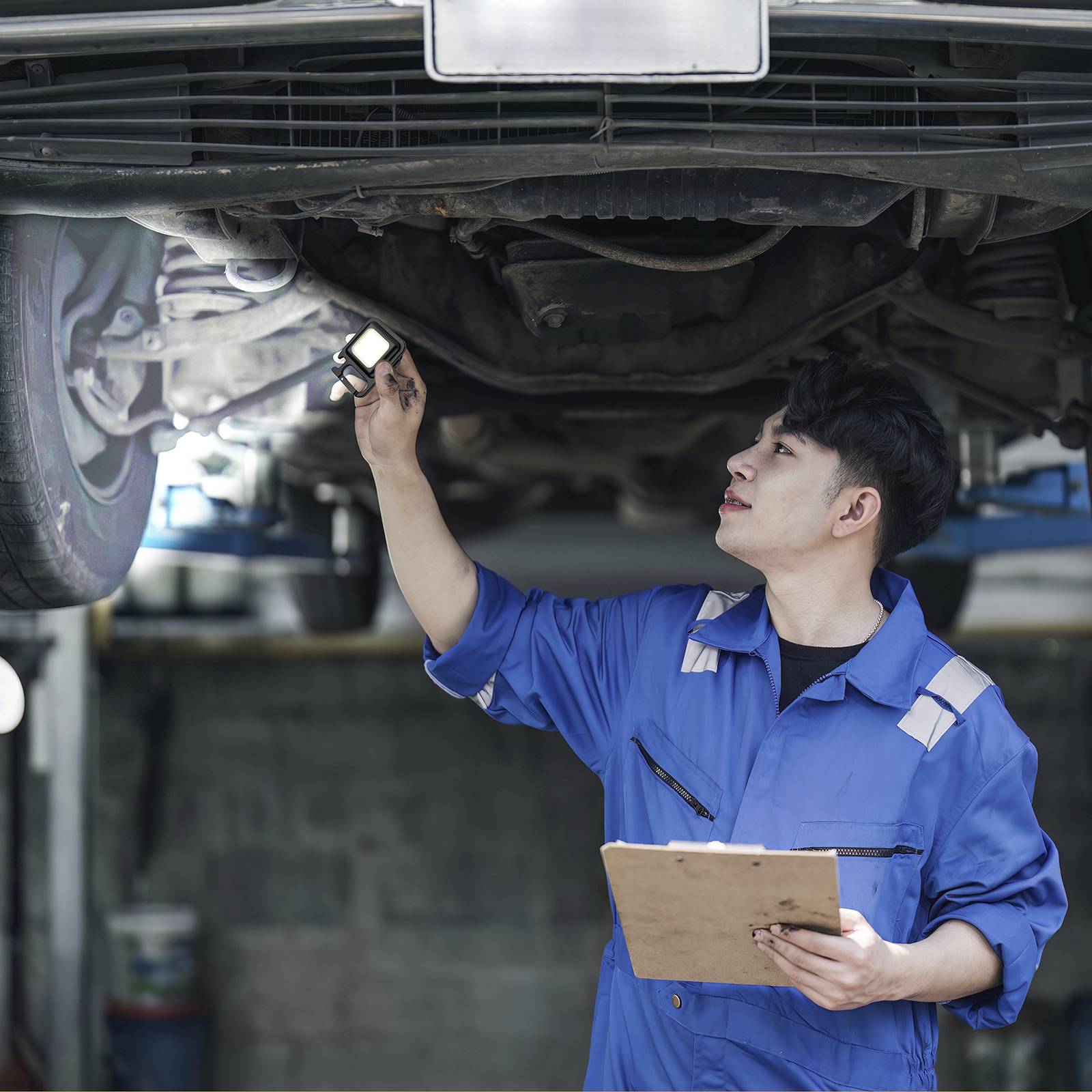 Monteur in blauwe werkkleding inspecteert een auto van onderen in een garage terwijl hij aantekeningen maakt op een klembord.