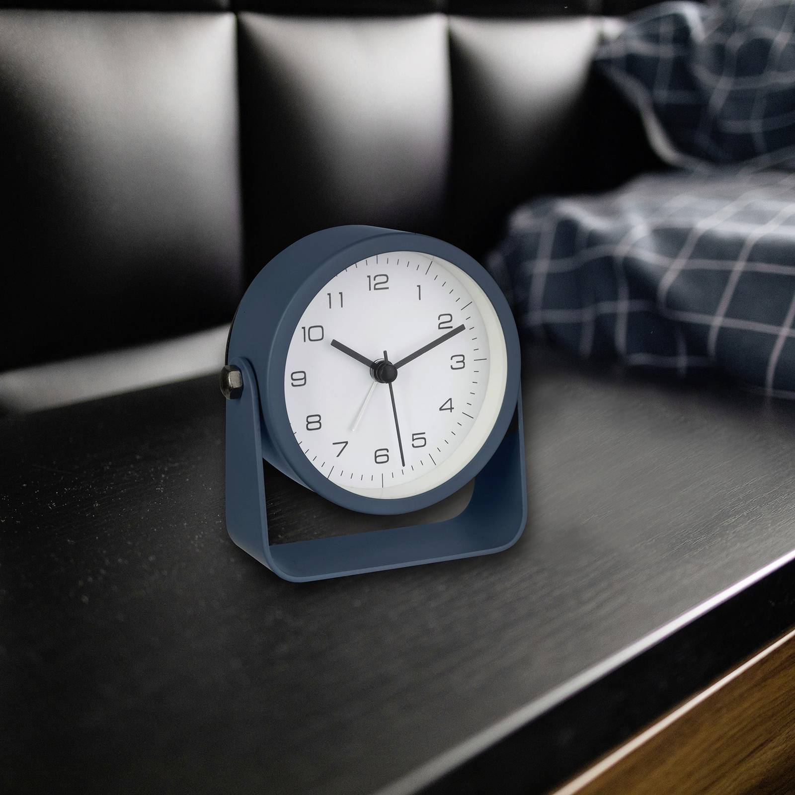 Een blauwe wekker toont 10:08 uur aan. Hij staat op een donkere houten tafel naast een bed met een geruit kussensloop.