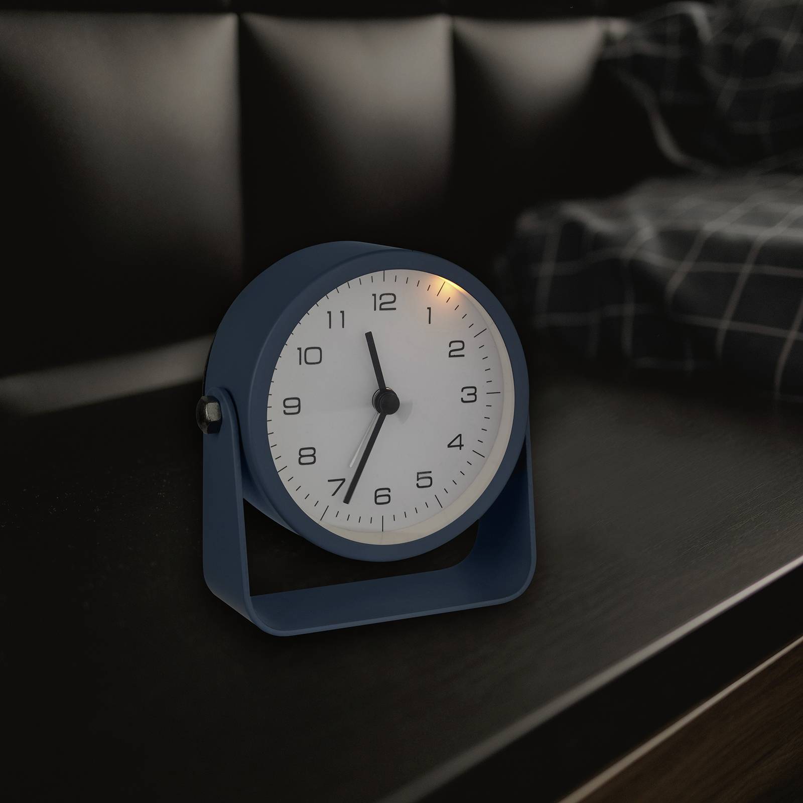 Een blauwe wekker toont 7:10 uur en staat in een donkere ruimte op een tafel naast een geruit dekbed.