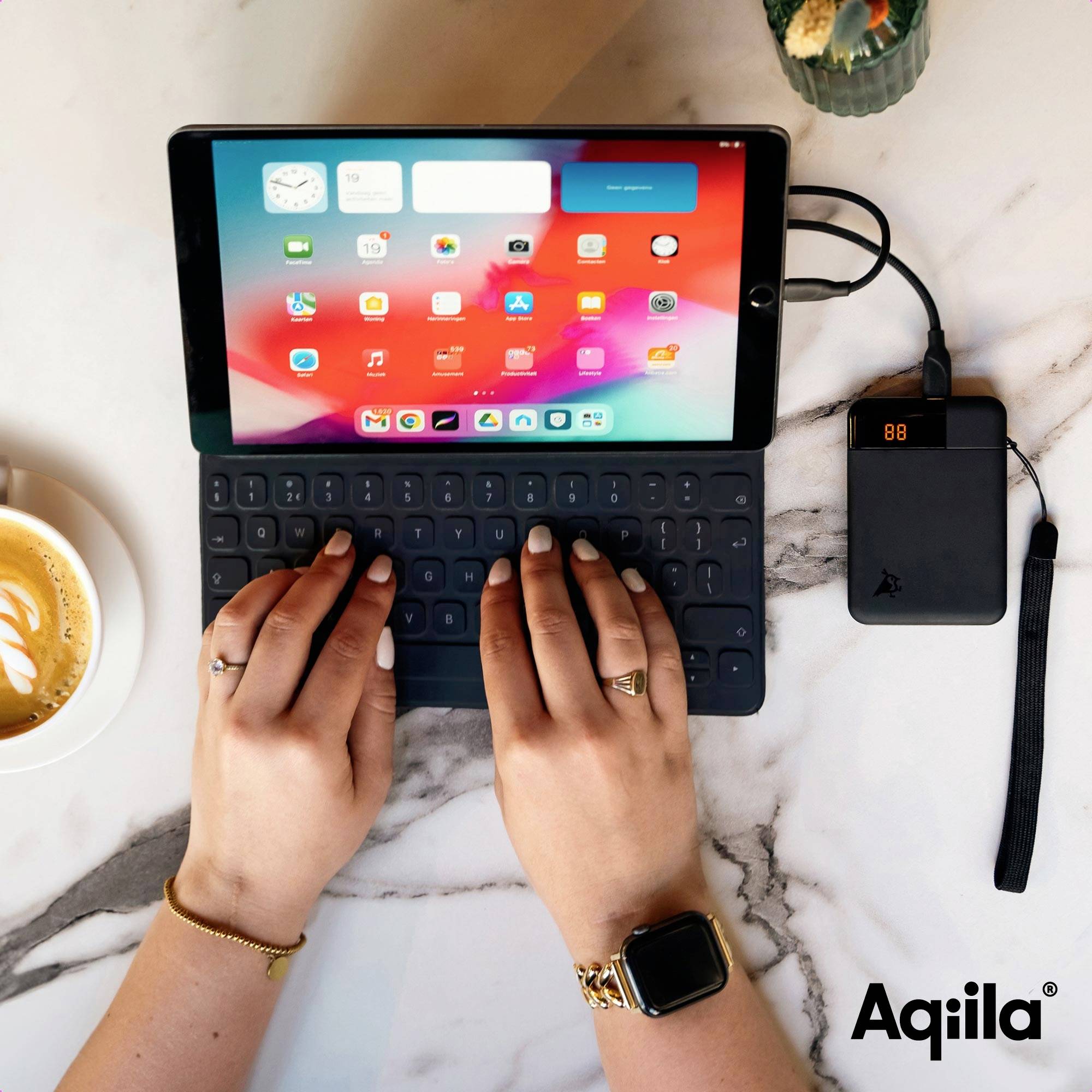 Een persoon typt op een tablet met een aangesloten toetsenbord. Een oplader is aangesloten. Rechtsonder is het 'Aqiila' logo zichtbaar.