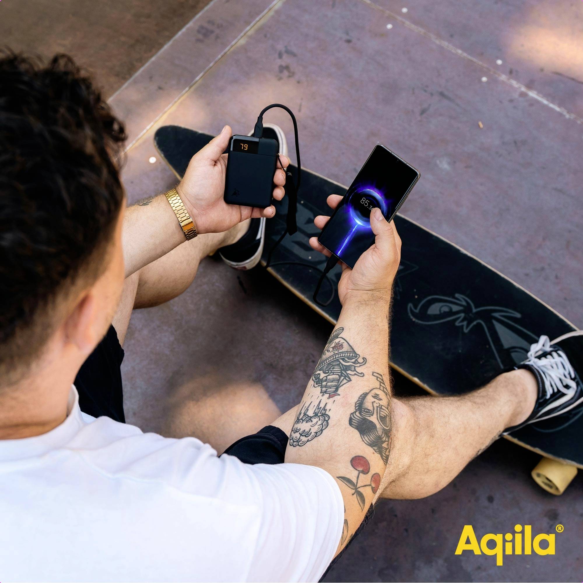 Een man zit op een skateboard en laadt zijn smartphone op met een powerbank. Hij heeft getatoeëerde armen. Op de achtergrond is de grond zichtbaar.