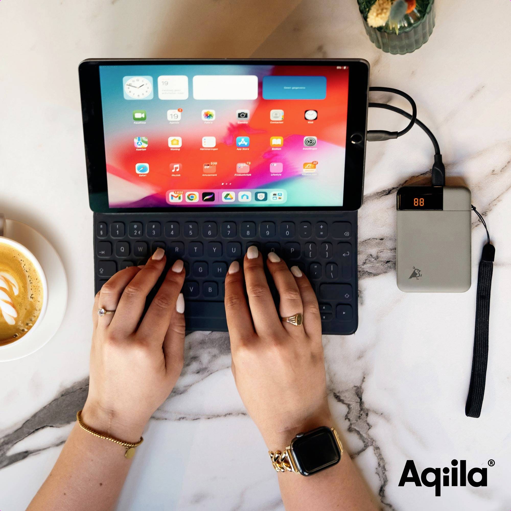 Een persoon typt op een tablet met een toetsenbord. Rechts daarnaast laadt een powerbank het tablet op. Links staat een kopje koffie.