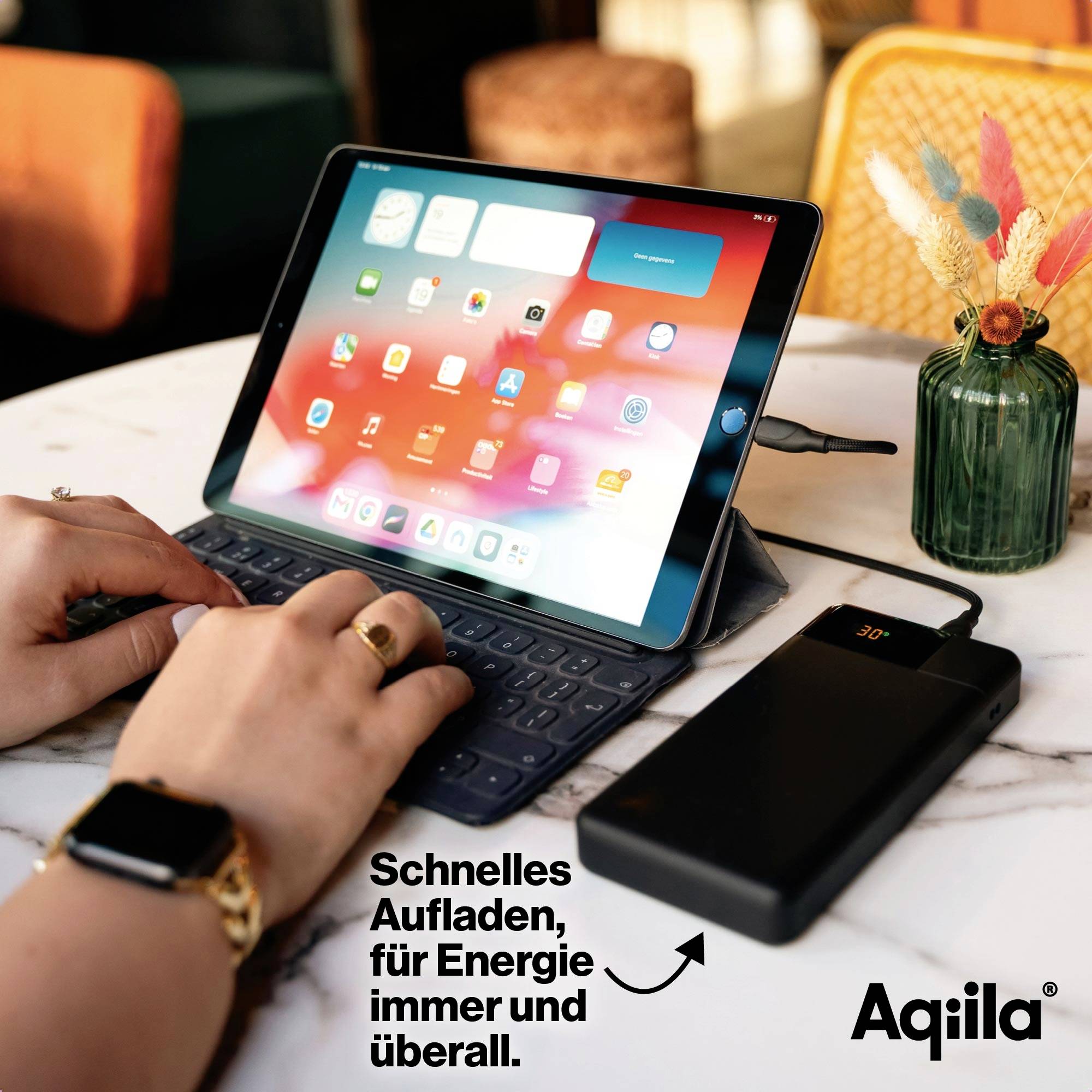 Tablet met toetsenbord op tafel, wordt opgeladen via powerbank. Naast het tablet steekt een hand. Tekst: 'Snel opladen, energie altijd en overal beschikbaar.'