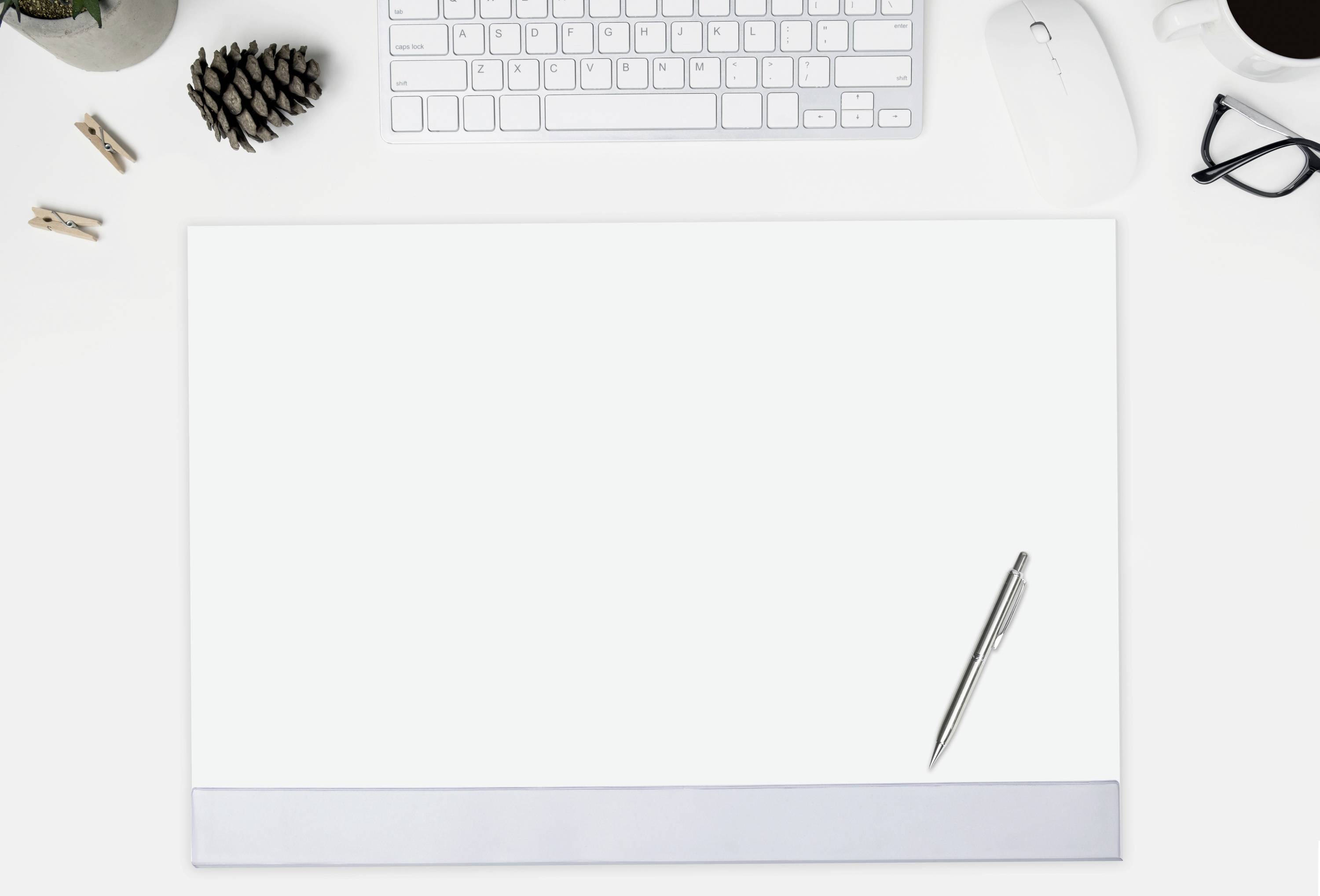 Een bureau met een computerkeyboard, muis, bril, koffiekopje en een leeg vel papier waarop een pen ligt.