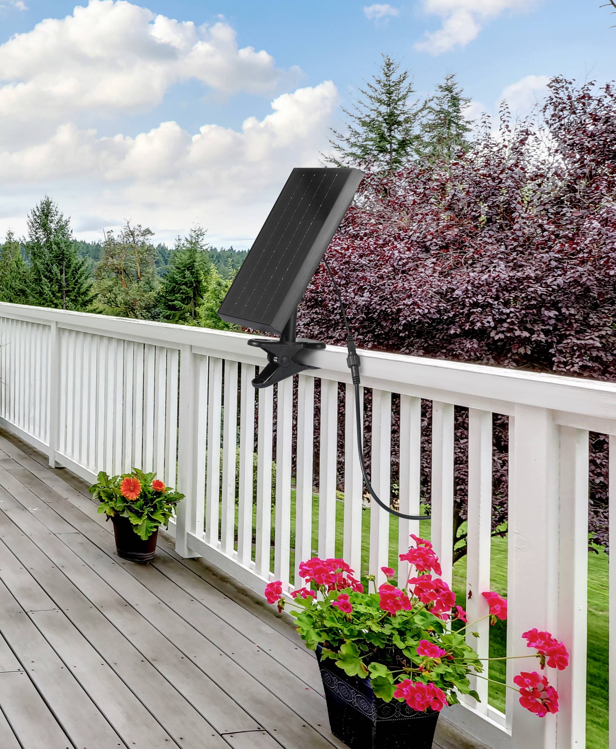Een balkonhek met een bevestigd zonnepaneel, bloeiende rode bloemen in potten eronder en een bewolkte lucht op de achtergrond.