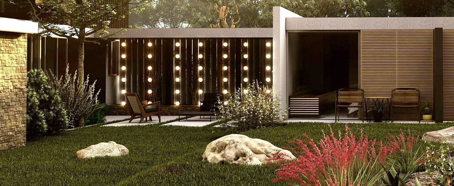 Een moderne tuin met gazon, decoratieve struiken en rotsen. Op de achtergrond een verlicht terras met houten meubels en planten.