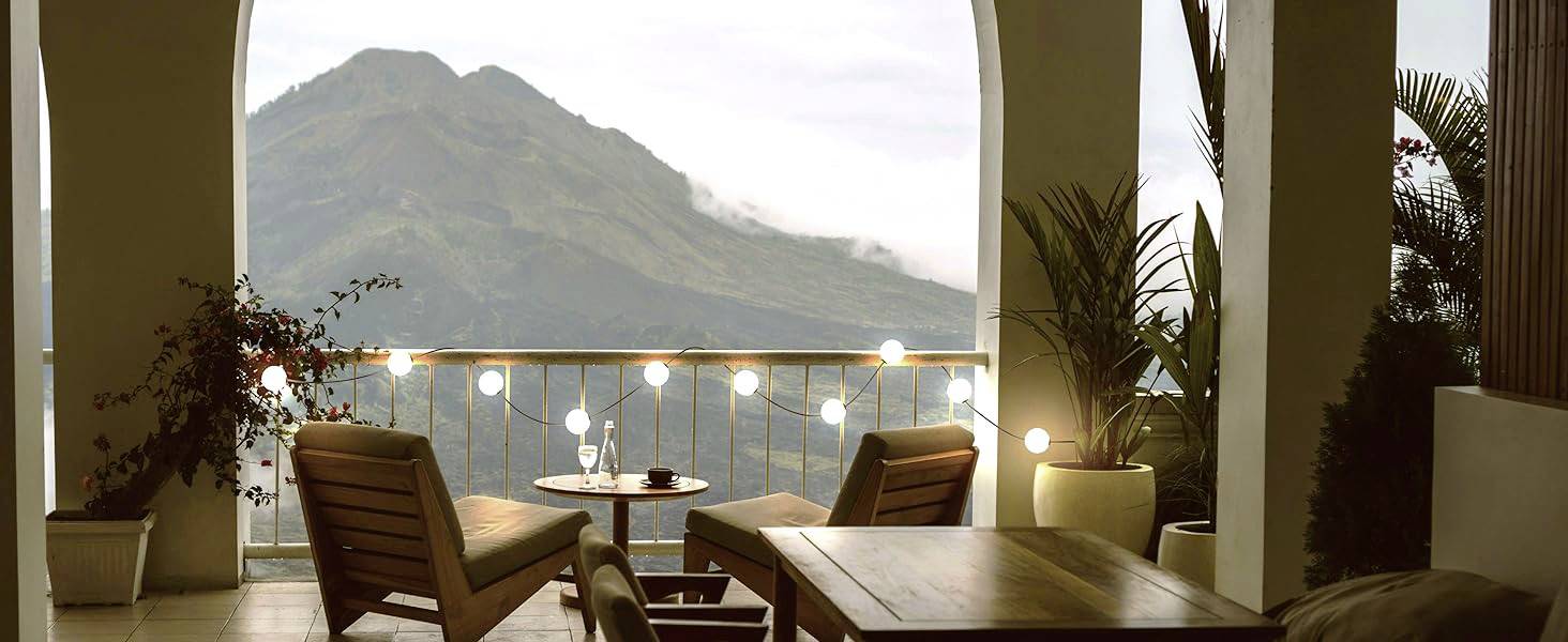 Een balkon met twee houten stoelen en een tafel biedt uitzicht op een met bomen bedekte berg op de achtergrond. Lichtjes sieren de balustrade.