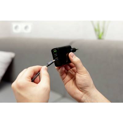 Een persoon houdt een oplader met meerdere USB-poorten vast en steekt een kabel in. Op de achtergrond is een bank zichtbaar.