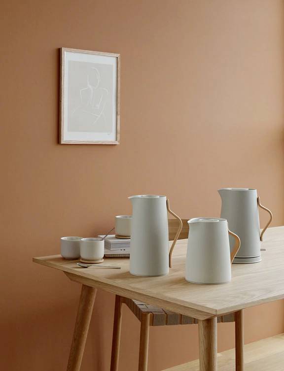 Een minimalistisch eetkamerinterieur met drie witte kannen en kopjes op een houten tafel voor een warme, oranje muur met een schilderij.