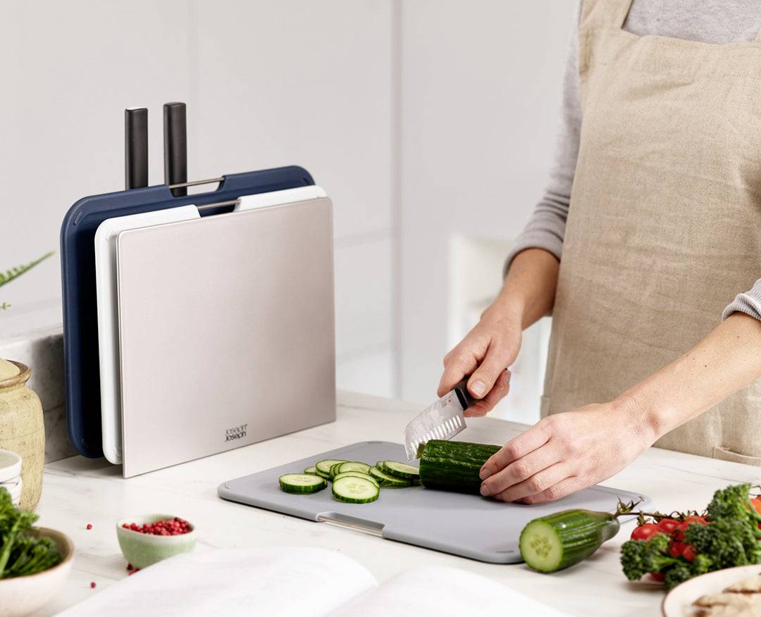 Een persoon snijdt komkommers op een snijplank in een moderne keuken. Ernaast staat een set met meerdere snijplanken.