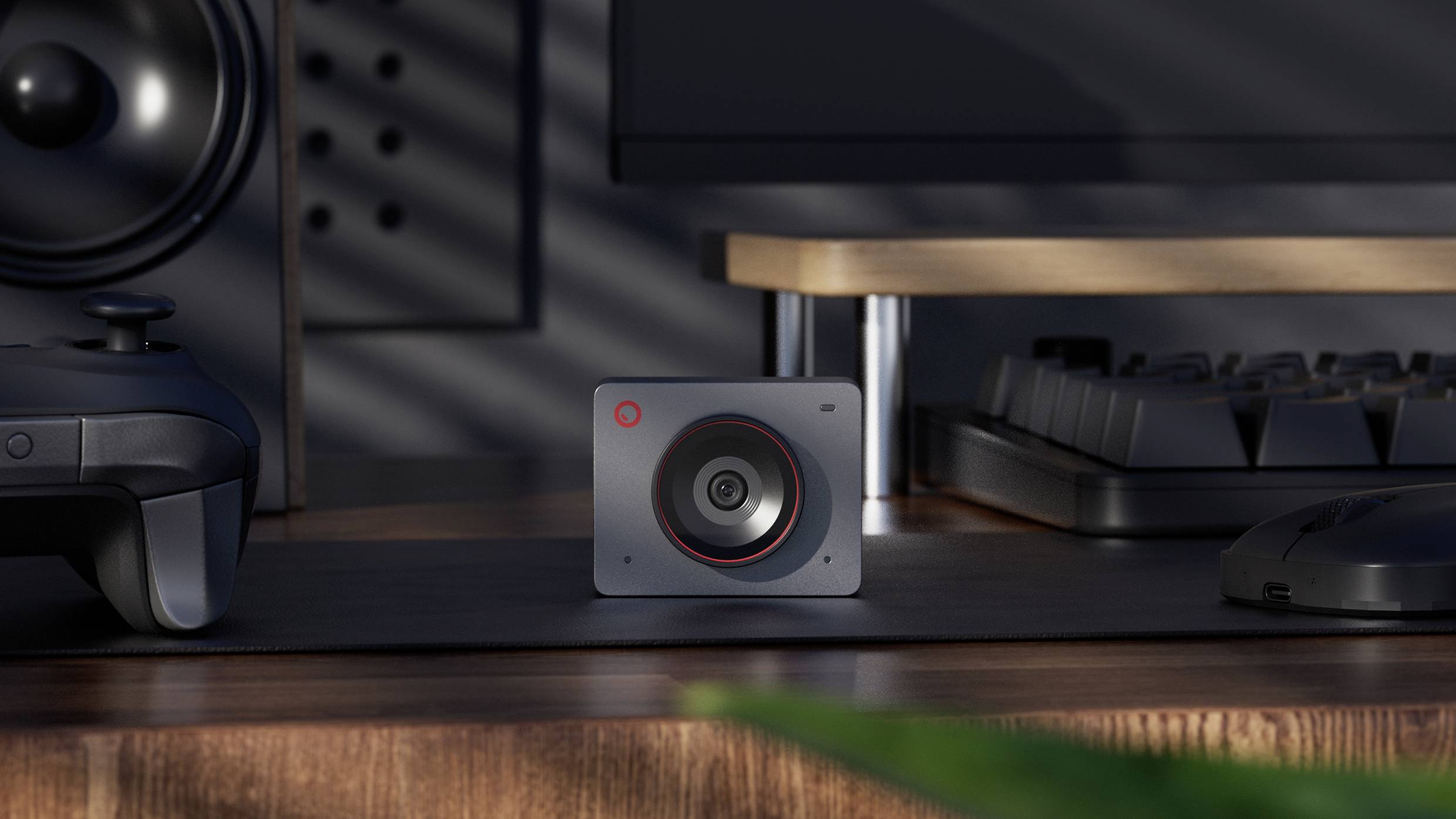 Een moderne luidspreker met een rode ring op een houten bureau, omringd door computeraccessoires en een grijze monitor op de achtergrond.