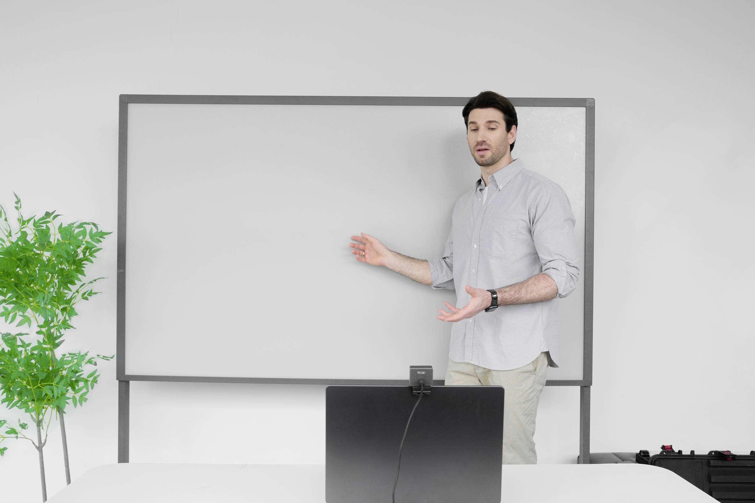 Een man staat voor een leeg whiteboard en houdt een presentatie. Voor hem staat een laptop. Links in het beeld staat een groene plant.