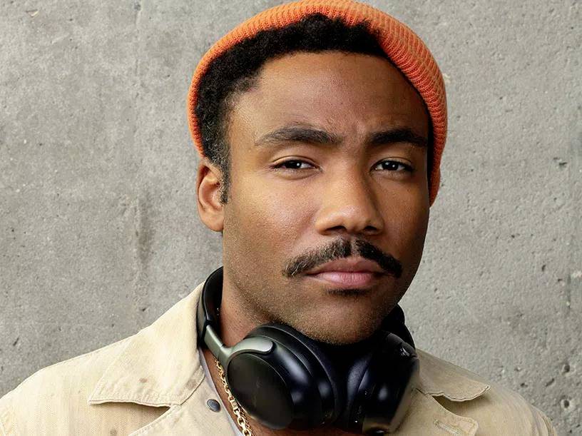 Ik weet niet wie deze persoon is. Een man met een oranje beanie, een snor en koptelefoons om zijn nek kijkt in de camera. De achtergrond is een grijze betonnen muur.
