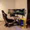 'Een racesimulator met stuur en meerdere monitors, toont een racespel. Op de achtergrond staat een verlichte computer.'