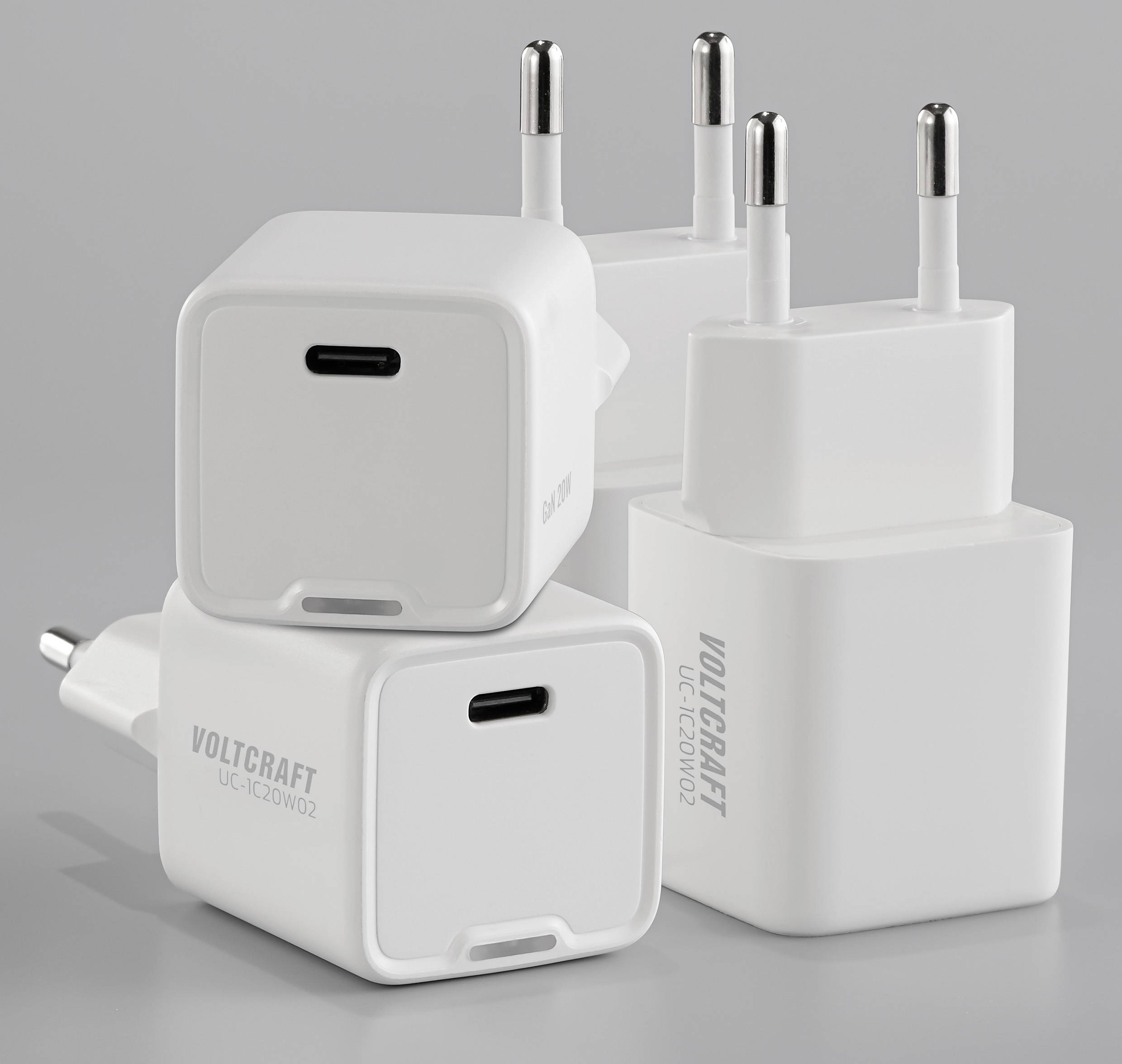 Vier witte USB-opladers met stopcontactaansluitingen staan rechtop op een grijze ondergrond; productnaam 'Voltcraft UC-120W02' zichtbaar.
