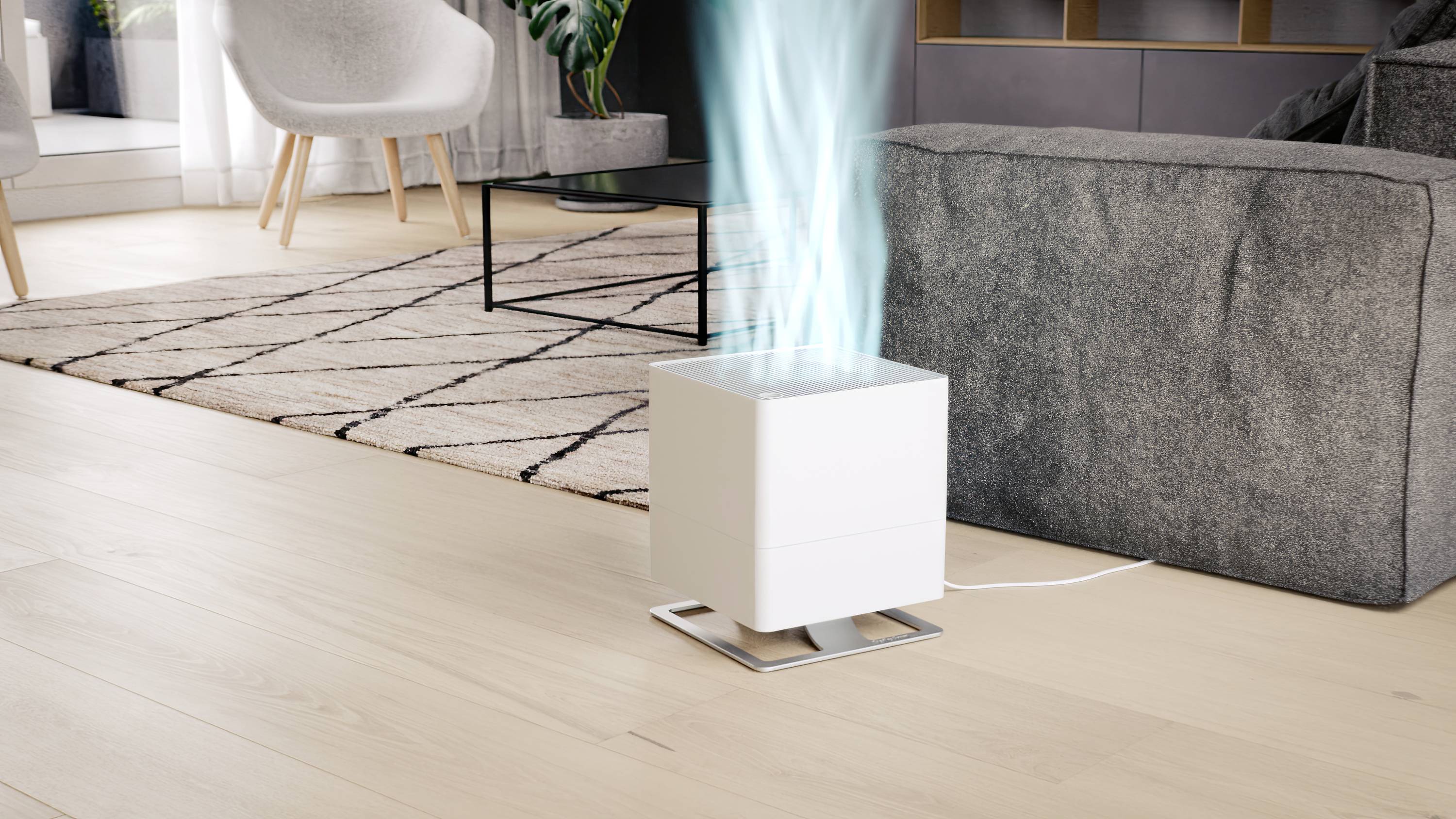 Een witte luchtreiniger staat op een houten vloer in een woonkamer en blaast zichtbaar schone lucht naar boven. Op de achtergrond een bank.