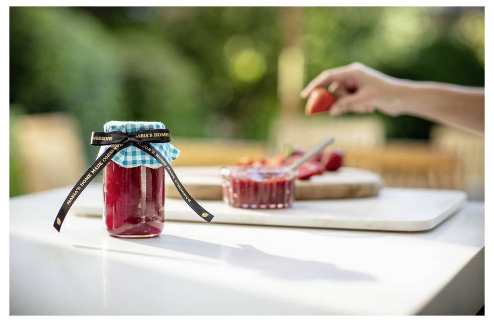 Een pot rode jam met een geruite stof en lint staat op tafel. Een hand houdt een aardbei vast boven een kom met aardbeien op de achtergrond.