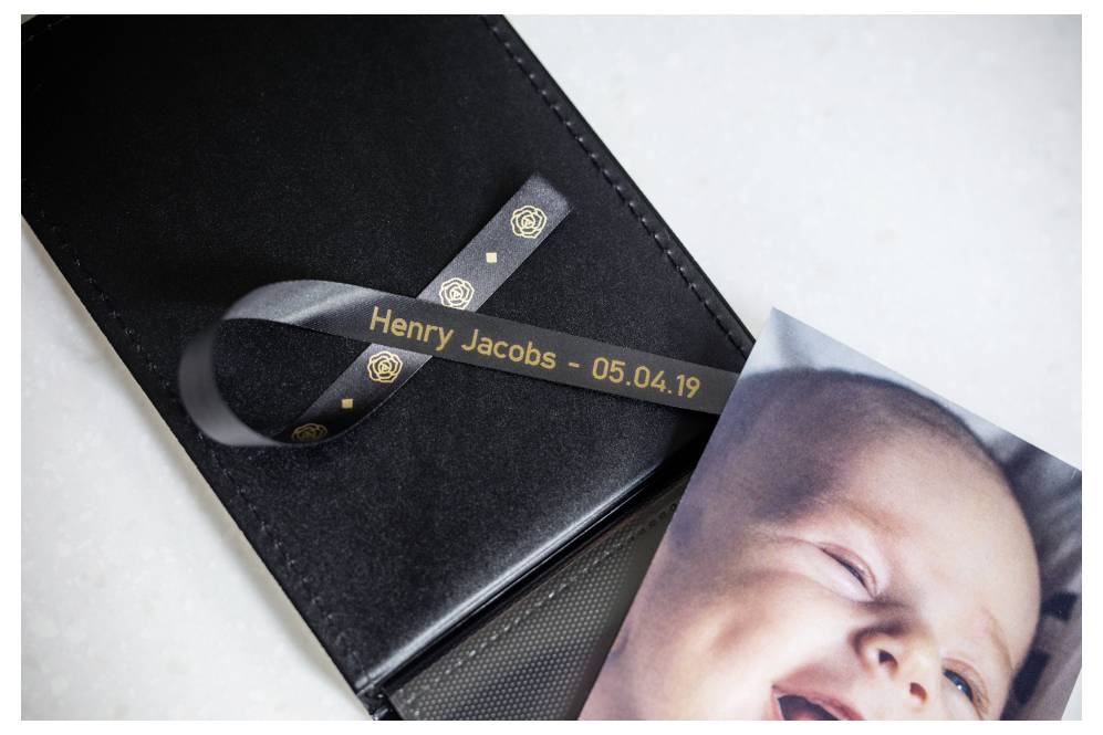 Een babyfoto met een zwart lint met de inscriptie 'Henry Jacobs - 05.04.19' geplaatst op een zwart leren portefeuille, wat lijkt te duiden op een herdenkingseerbetoon.