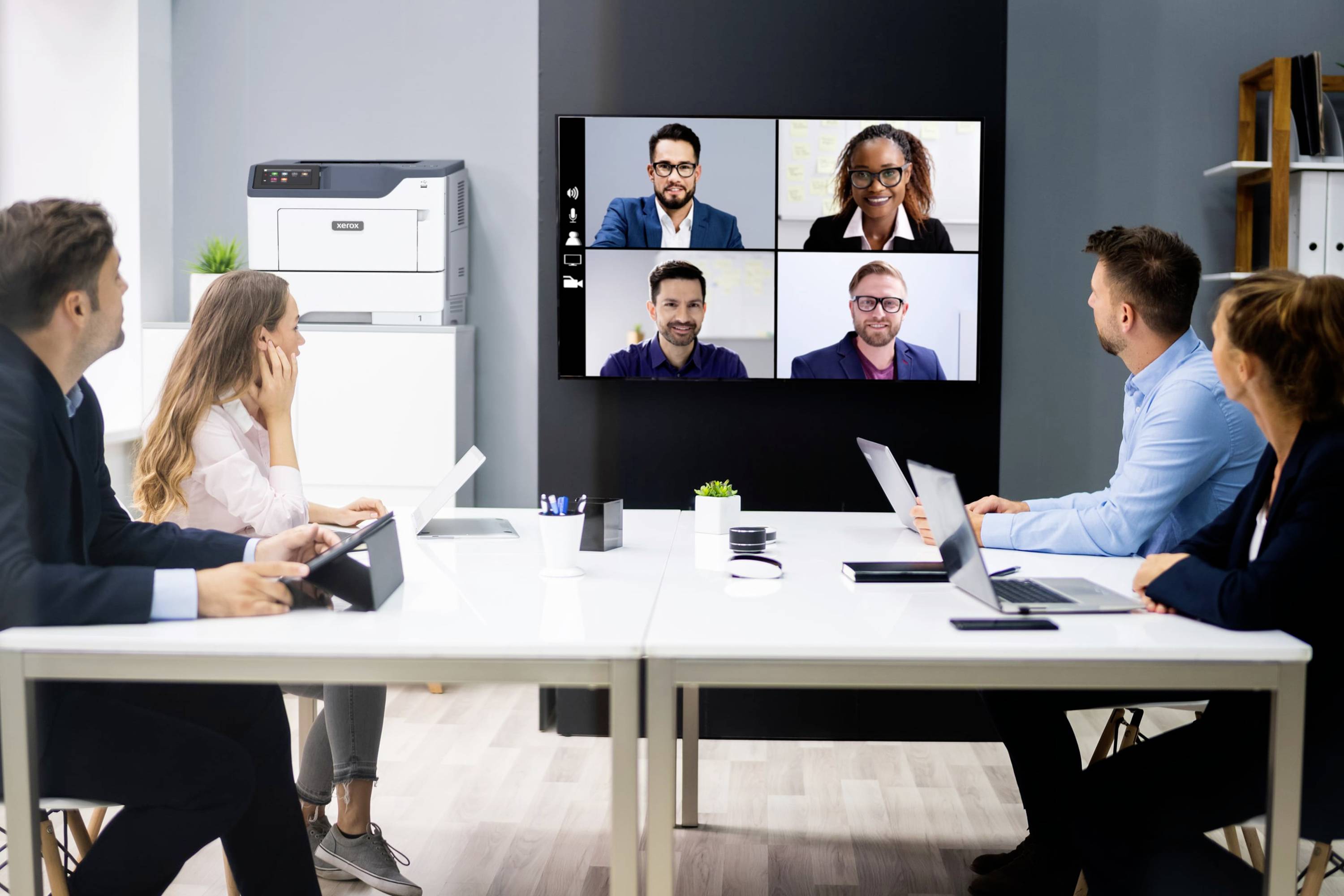 Zes personen in een vergaderruimte nemen deel aan een virtuele vergadering. Vier personen zijn zichtbaar op een scherm aan de muur.