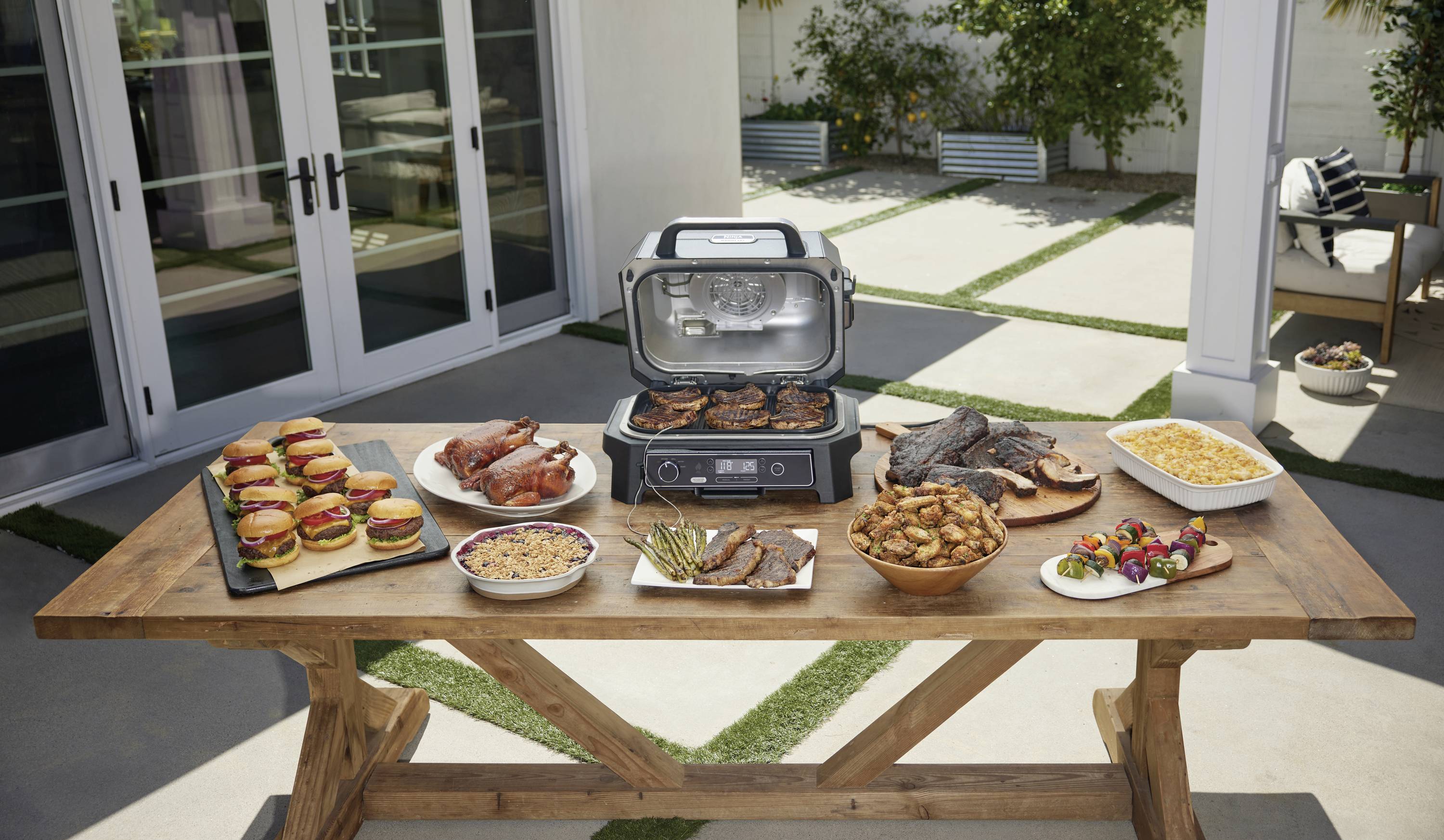 Een houten tafel buiten met een grill in het midden, omringd door borden met hamburgers, kip, groenten, vlees en bijgerechten.