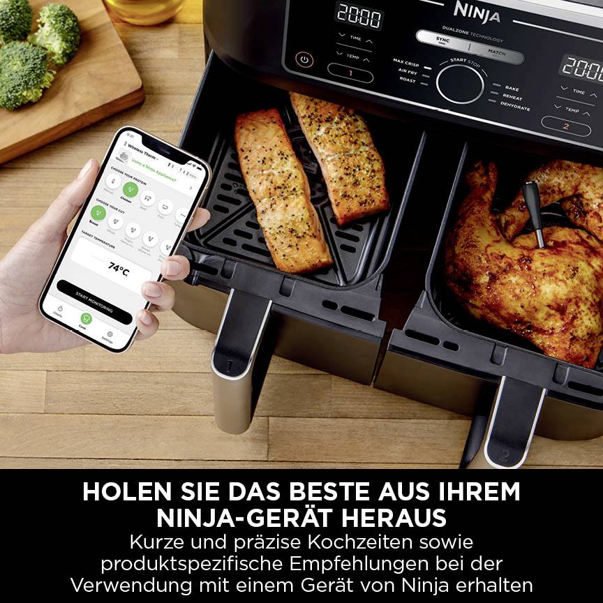 Een persoon houdt een smartphone vast met een app die 74 °C weergeeft, naast een tweevoudig geopende friteuse met eten erin.