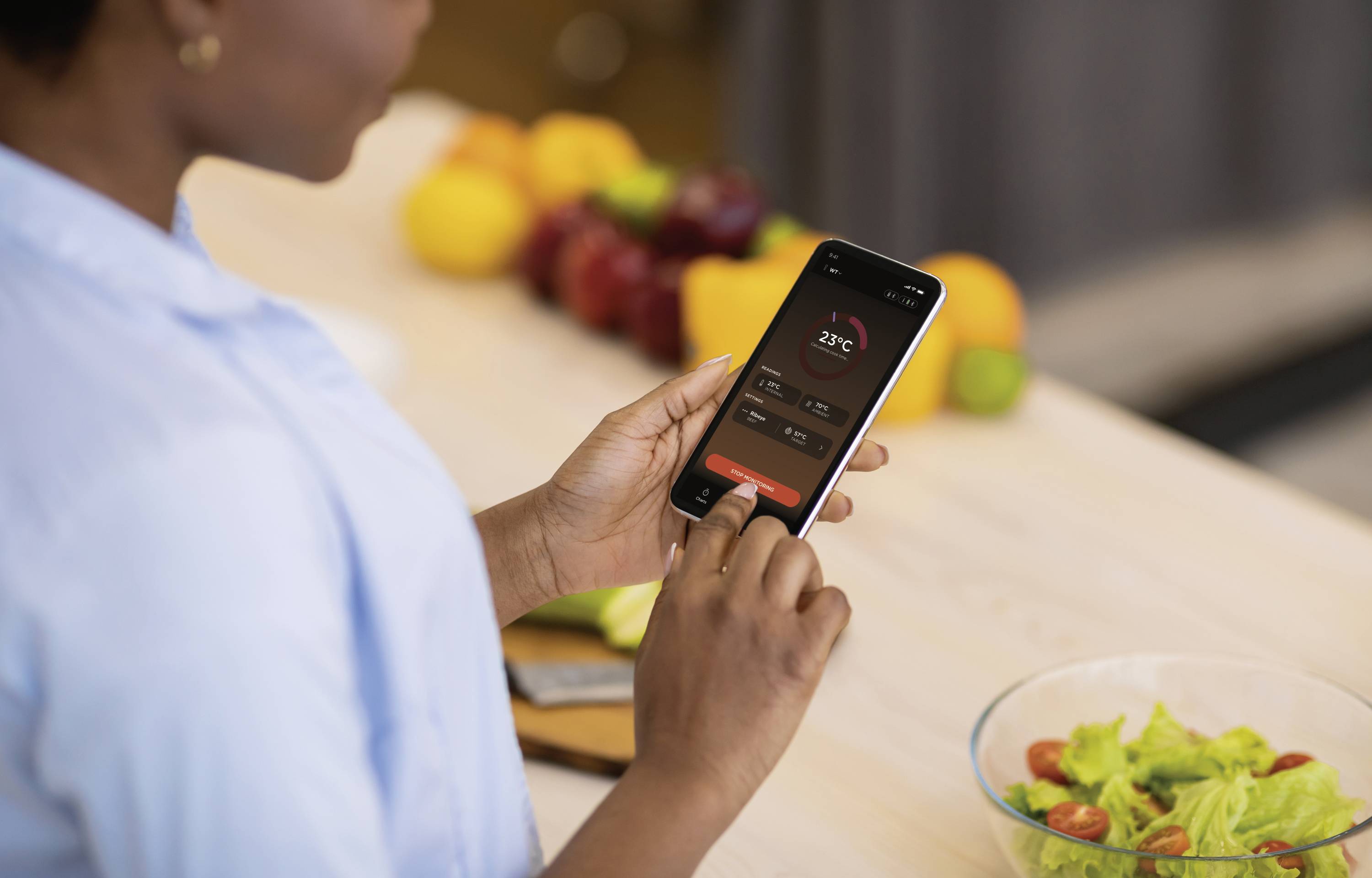 Een persoon gebruikt een app op een smartphone om een temperatuur van 23°C in te stellen. Naast de smartphone staat een kom met salade.