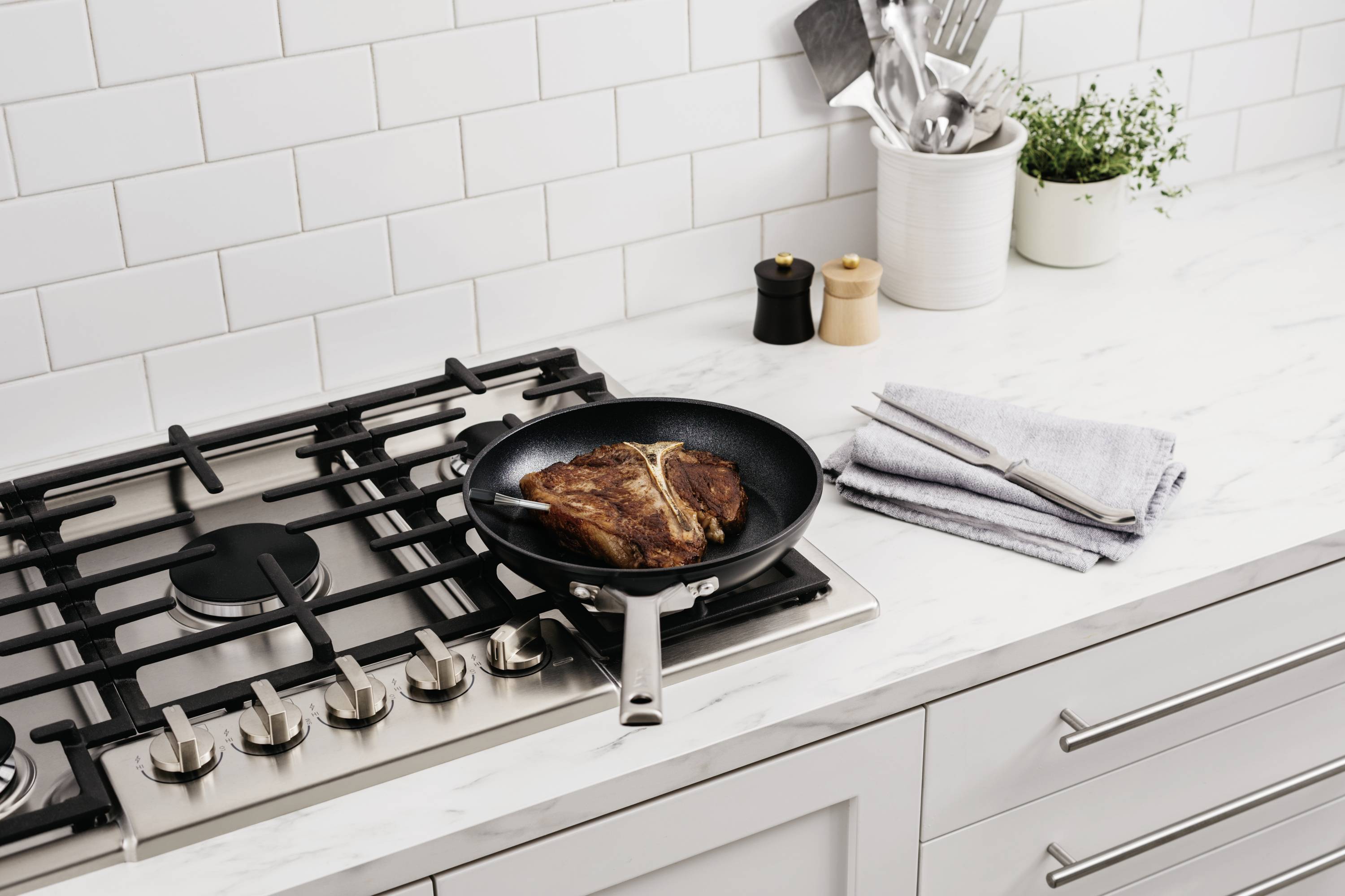 Een pan met gebakken vlees op een gaskookplaat in een moderne keuken, naast een handdoek en keukengereedschappen.