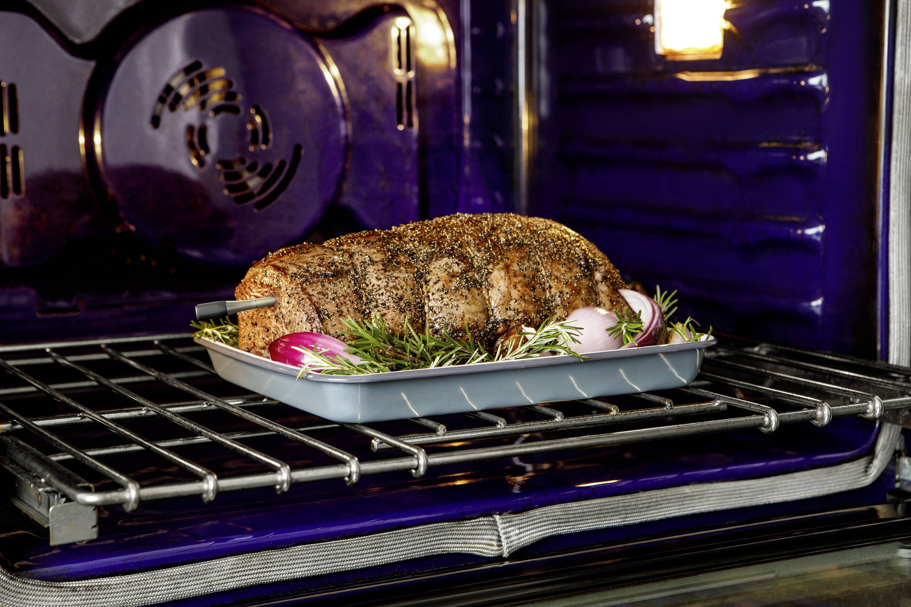 Een gekruide braadstuk ligt in een bakblik op een ovenrooster, omgeven door uien en rozemarijn. De oven staat aan.