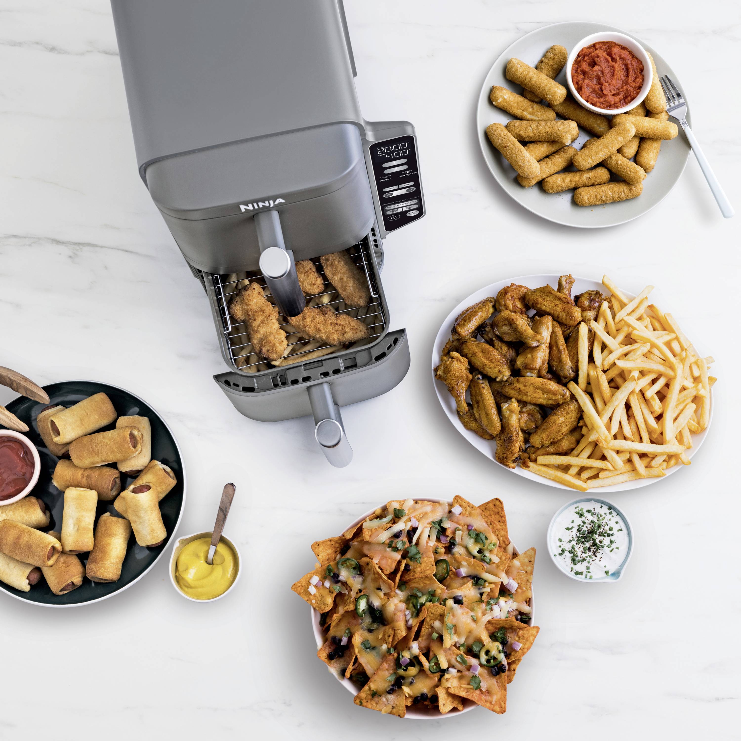 Een airfryer frituurt kipstroken. Op tafel liggen frietjes, chicken wings, lente-rolletjes, mozzarellasticks en beladen nacho's.