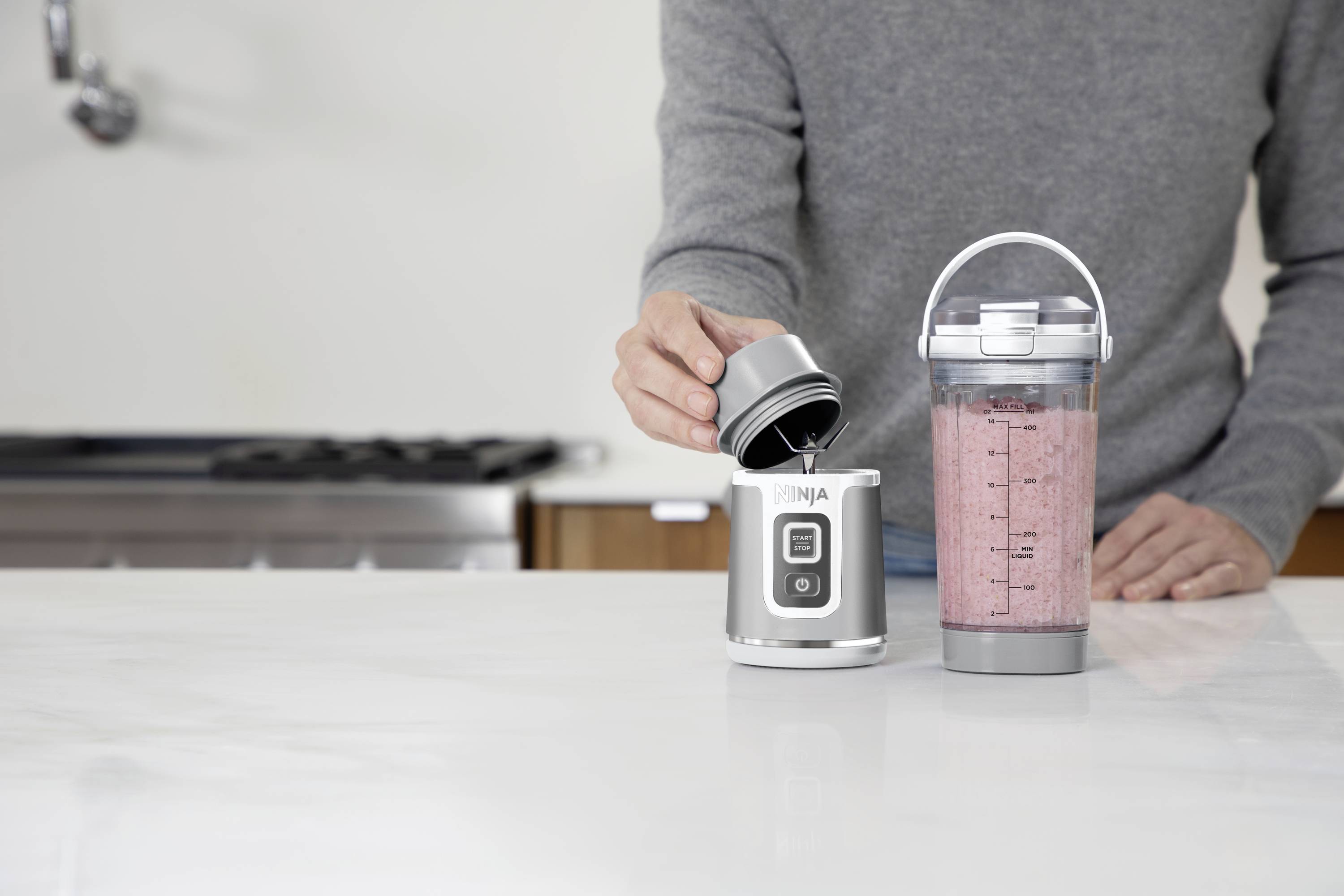 Een persoon giet vloeistof in een kleine blender. Naast de blender staat een gevulde shaker met een rood drankje. Keuken op de achtergrond.