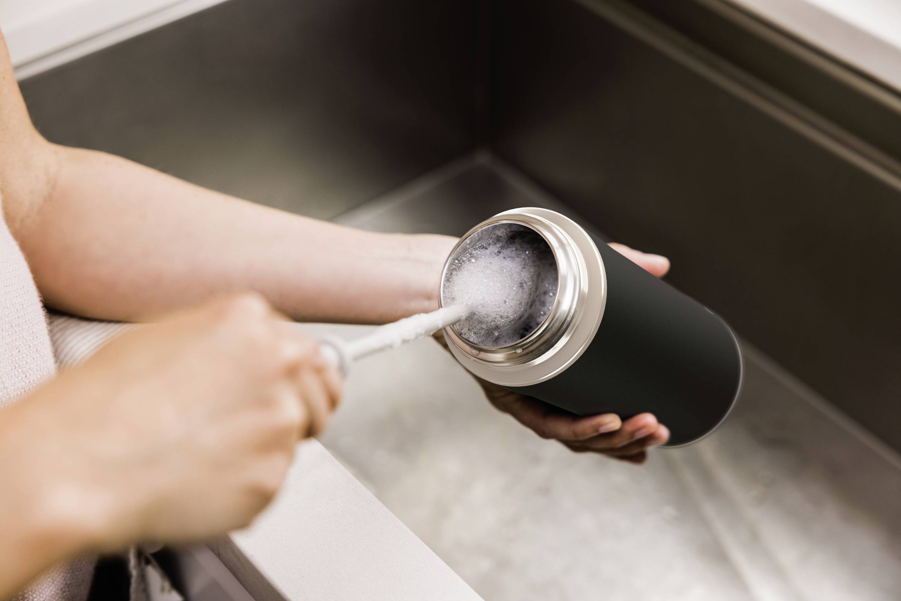 Persoon reinigt thermosfles met een borstel bij de wasbak. Water stroomt erin.