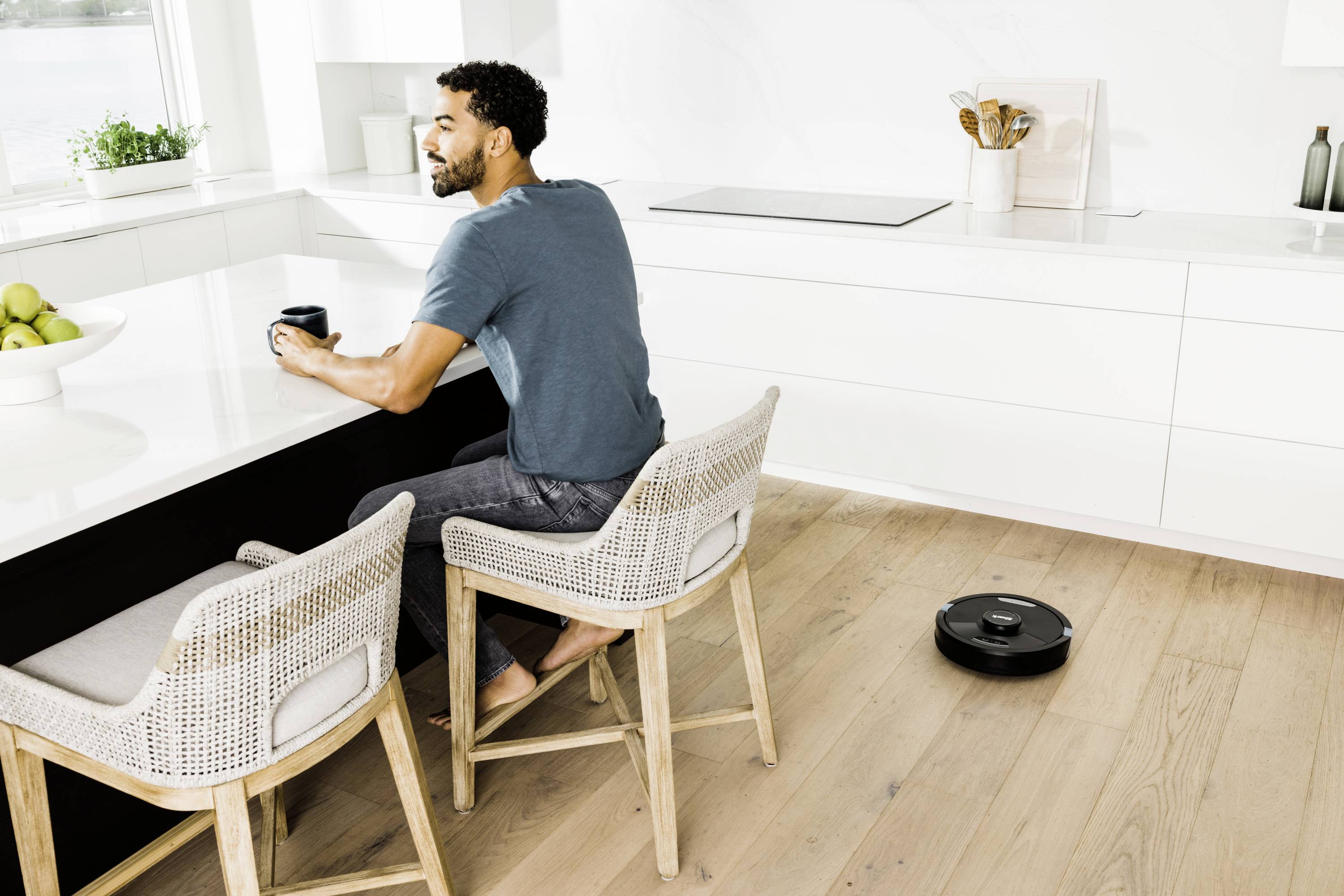 Een man zit aan de keukentafel met een mok, terwijl een robotstofzuiger de vloer schoonmaakt. Op de achtergrond is een moderne keuken te zien.
