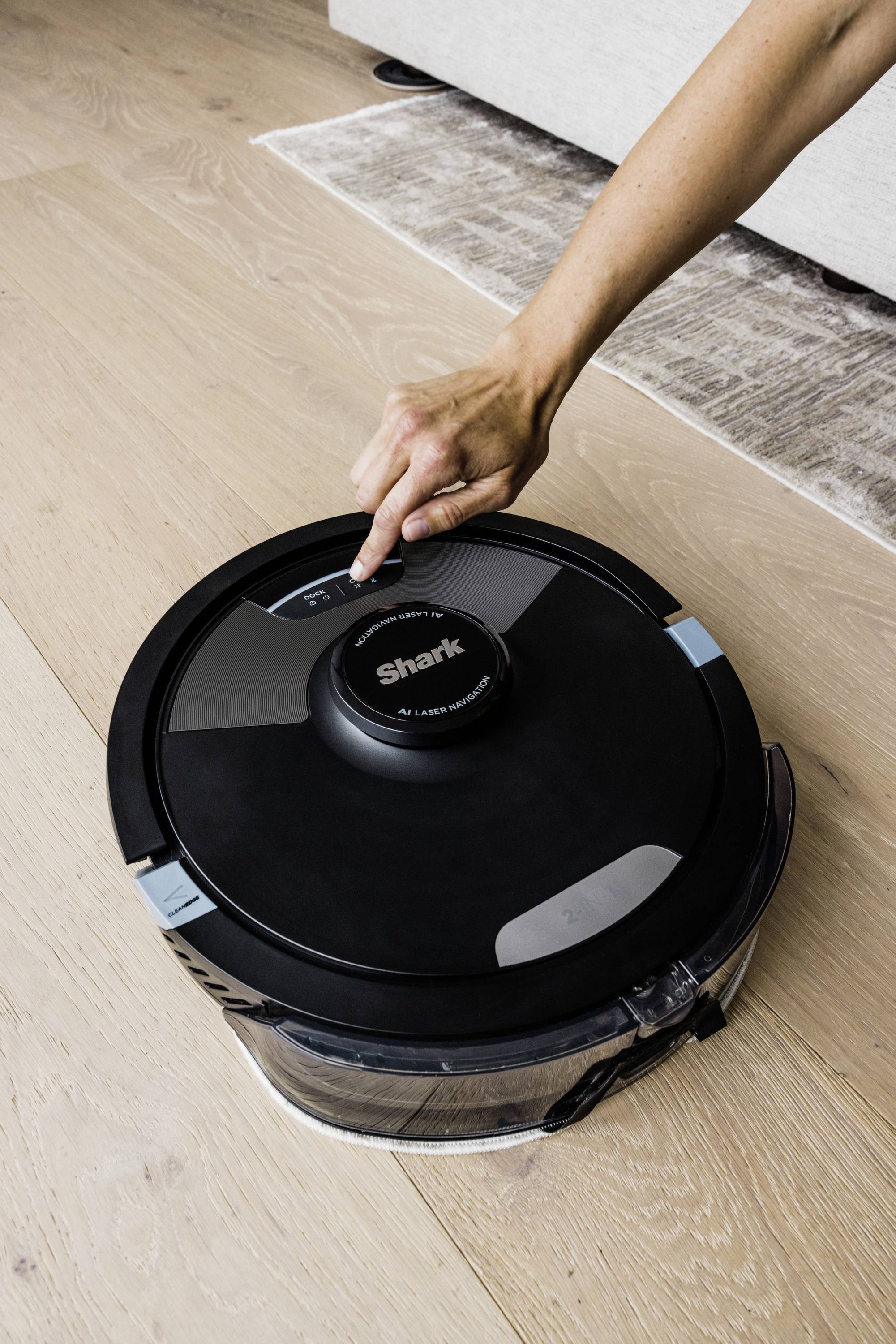 Een hand drukt op een ronde, zwarte stofzuigerrobot op een houten vloer. De robot is van het merk "Shark".