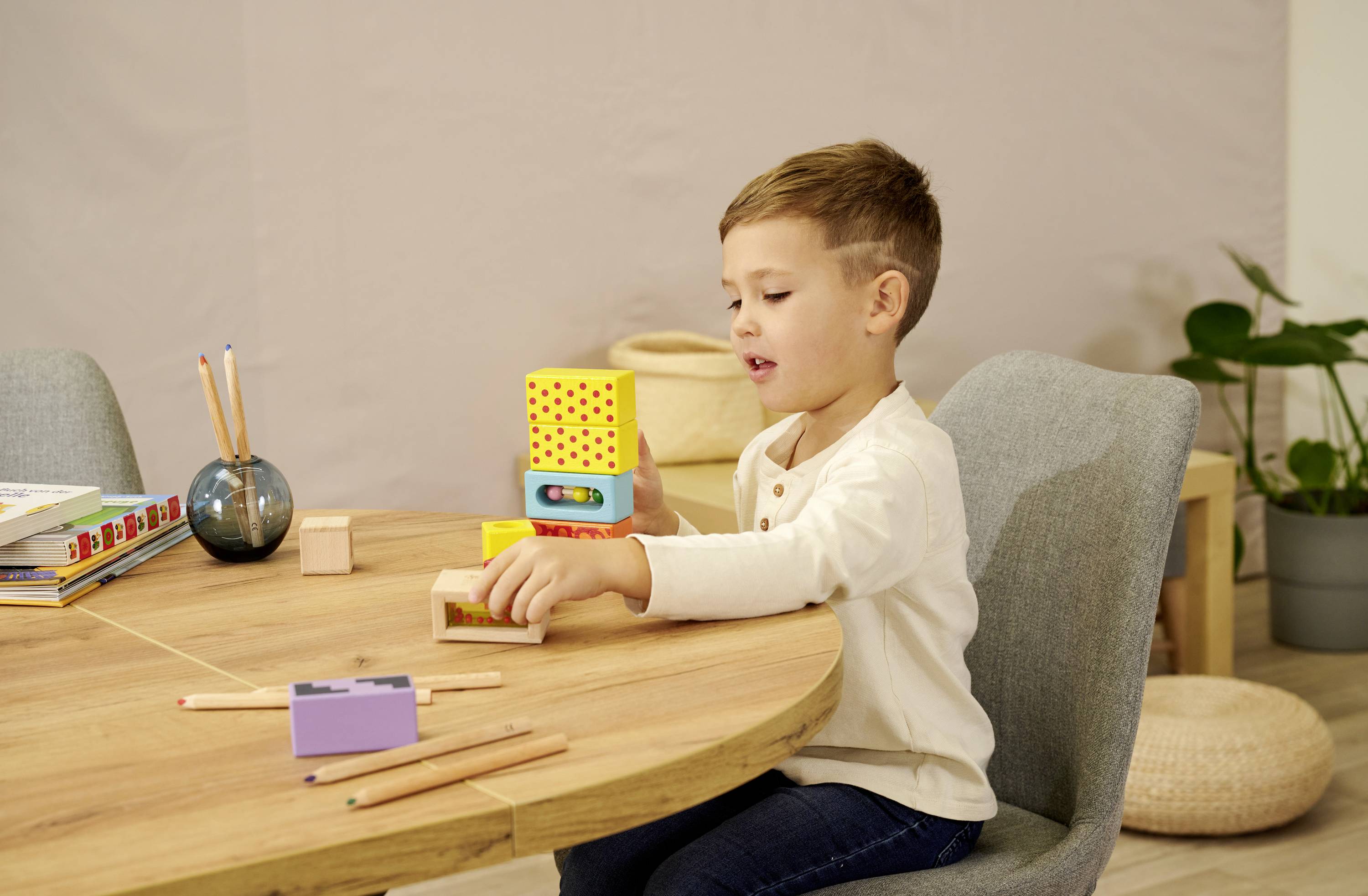 Een jongen zit aan een tafel en speelt met gekleurde houten blokken. Op de achtergrond zijn gekleurde potloden en boeken te zien.