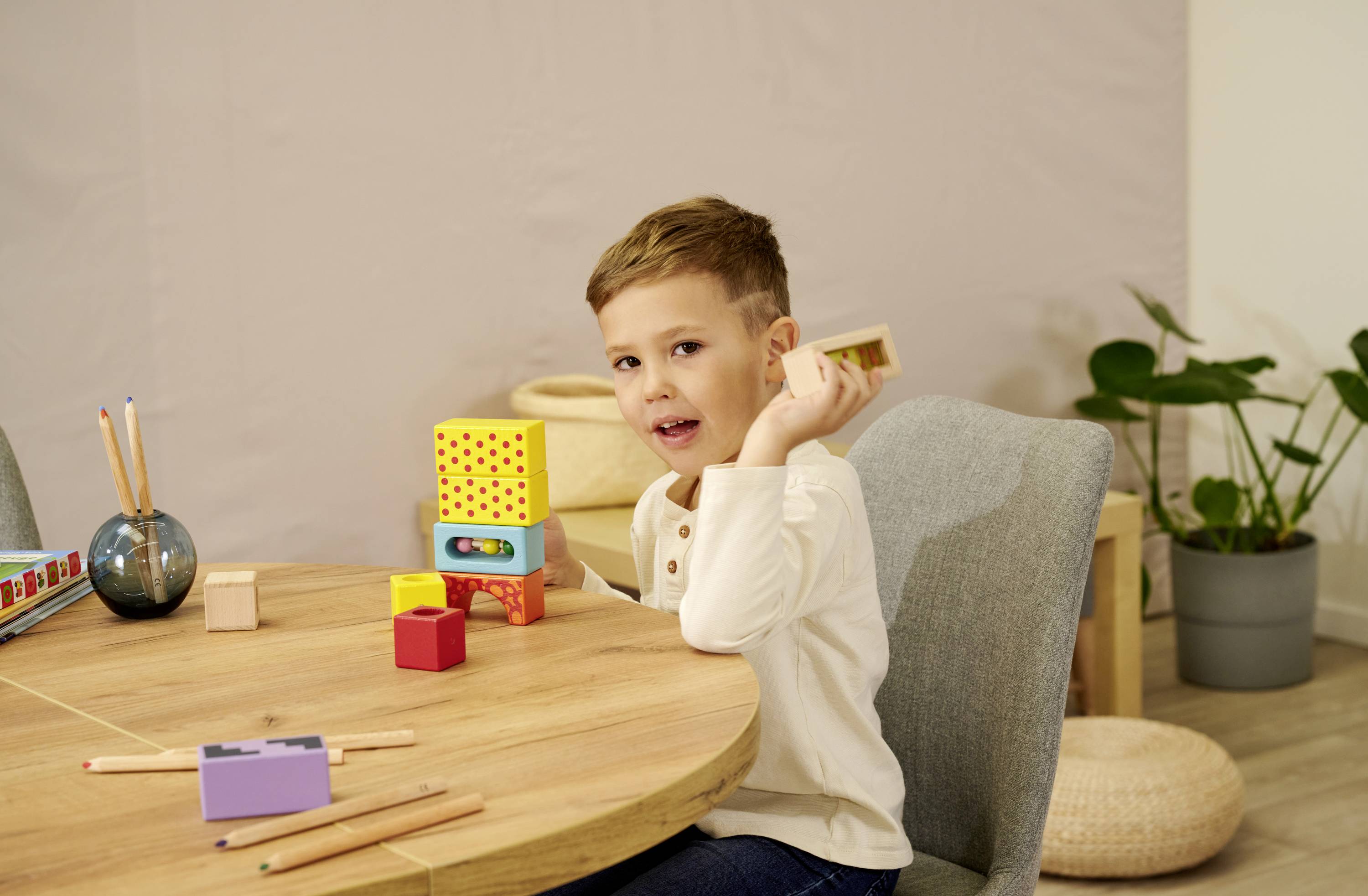Een kind zit aan een houten tafel en bouwt een toren van kleurrijke houten blokken. Op de achtergrond zijn planten en stoelen zichtbaar.