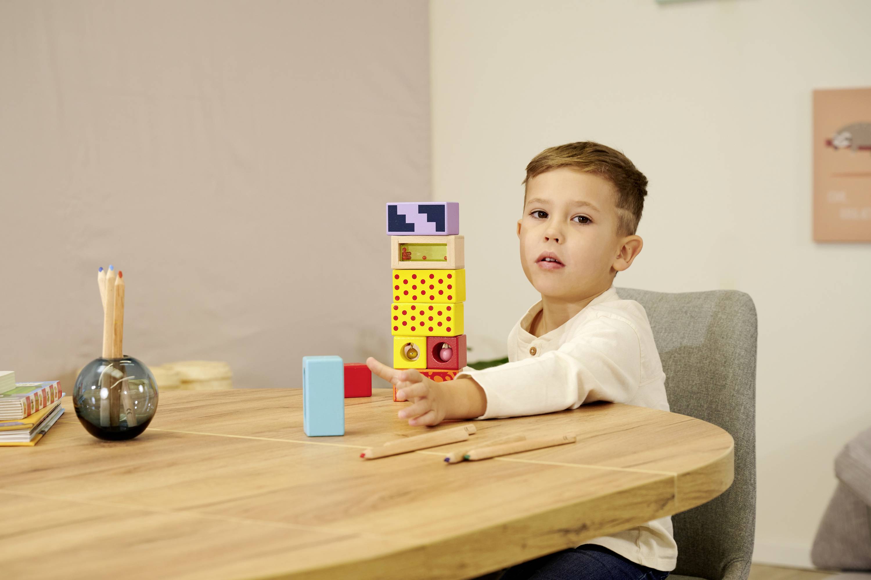 Een kind zit aan een tafel, speelt met kleurrijke bouwblokken en kijkt naar voren. Op de achtergrond zijn potloden en boeken.