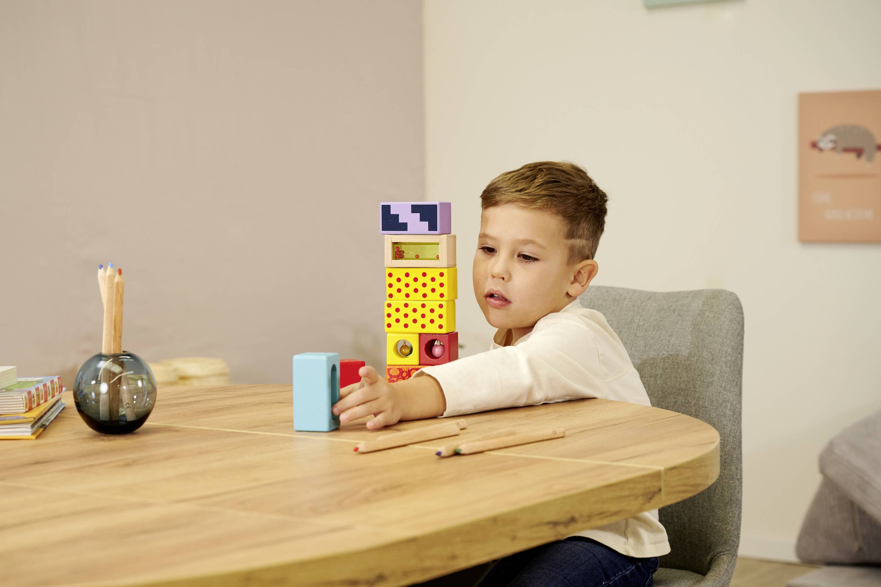 Een kind speelt met kleurrijke houten bouwblokken op een tafel. Op de achtergrond zijn een pennenbak en boeken te zien.