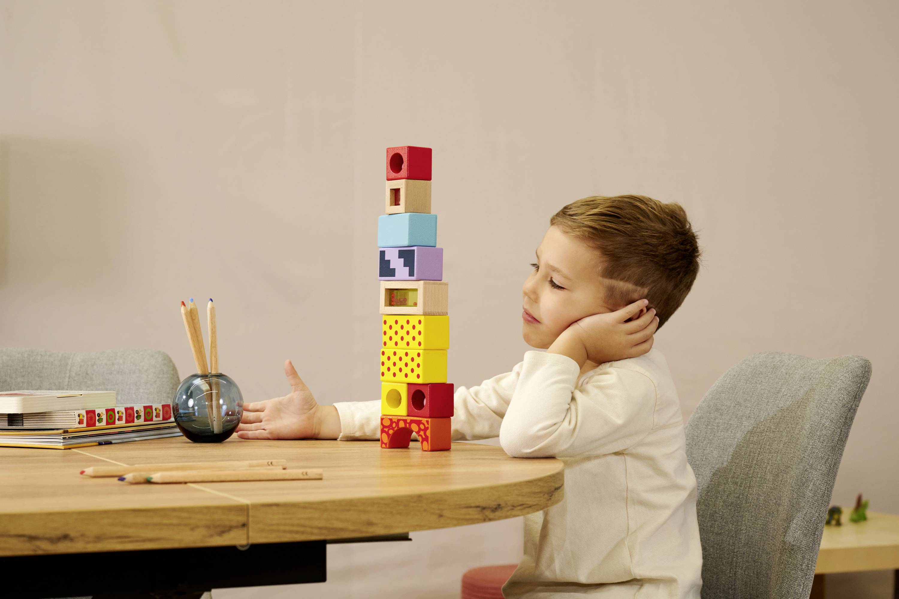 Een jongen zit aan een tafel en kijkt nadenkend naar een toren van kleurrijke blokken. Op tafel liggen potloden en een liniaal.