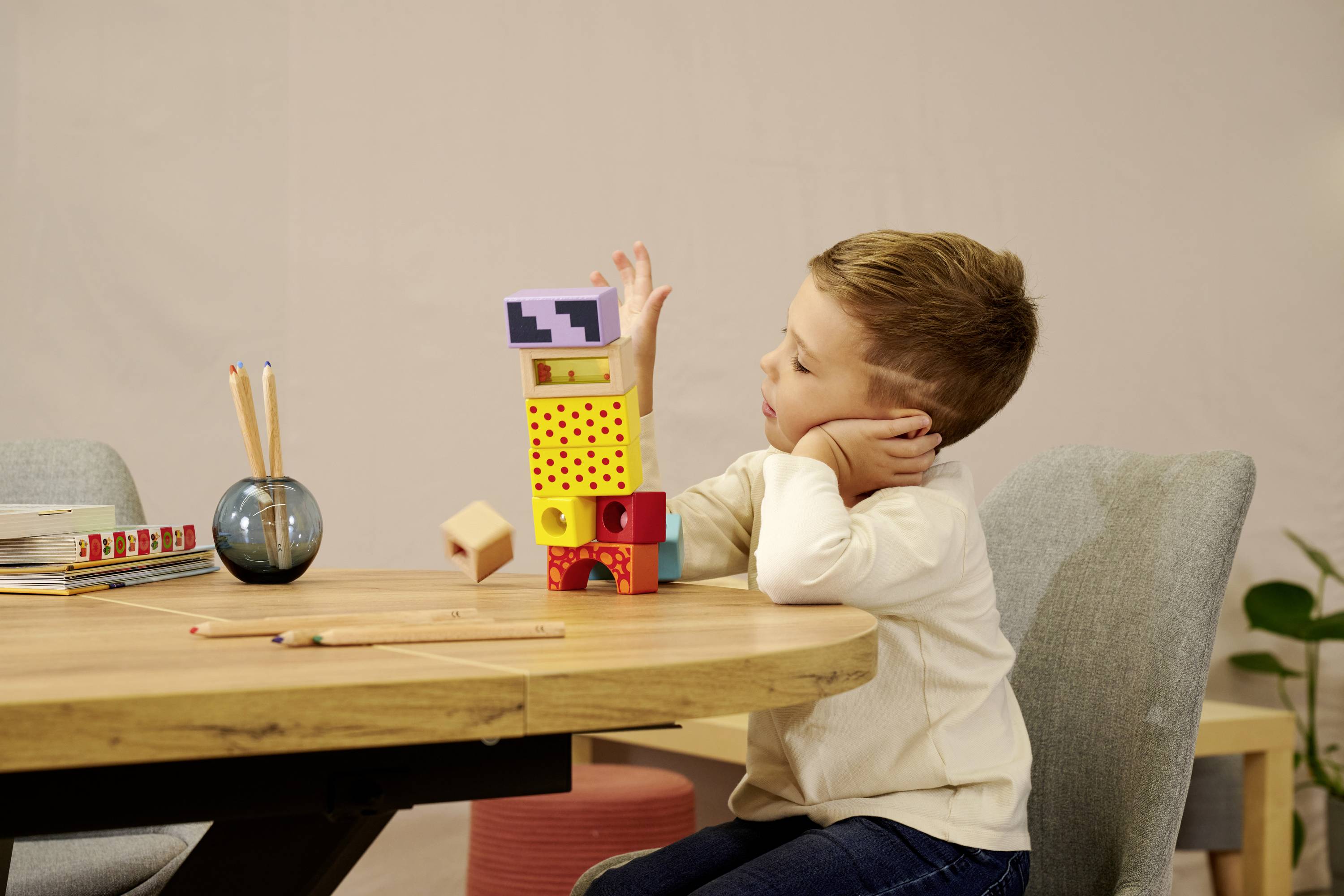 Een kind zit aan een tafel en bouwt met gekleurde houten blokken een toren. Naast het kind liggen kleurpotloden en een schrift.