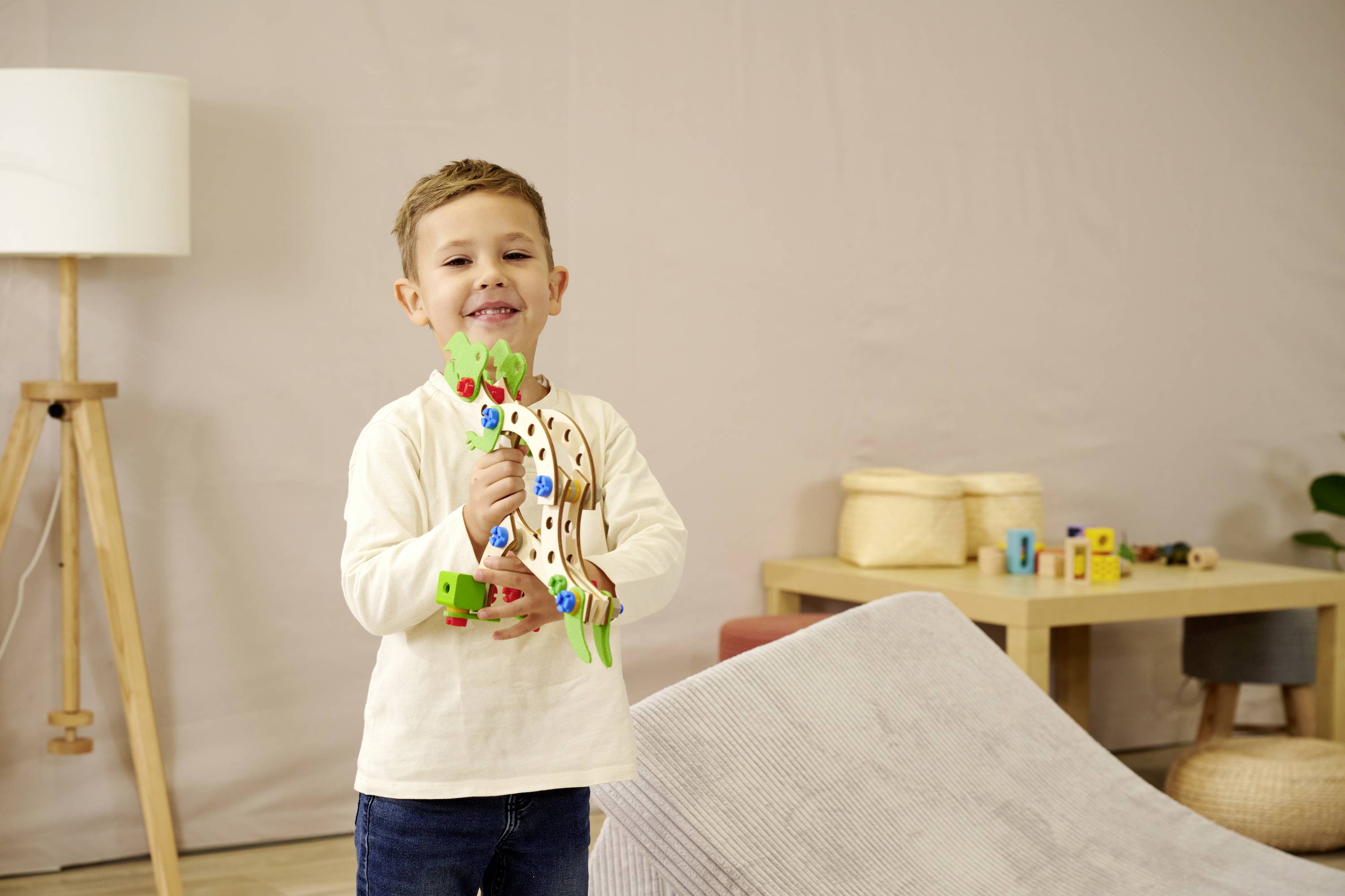 Een lachende jongen speelt in een woonkamer met een kleurrijk houten speelgoed. Op de achtergrond zijn een tafel en speelgoed zichtbaar.